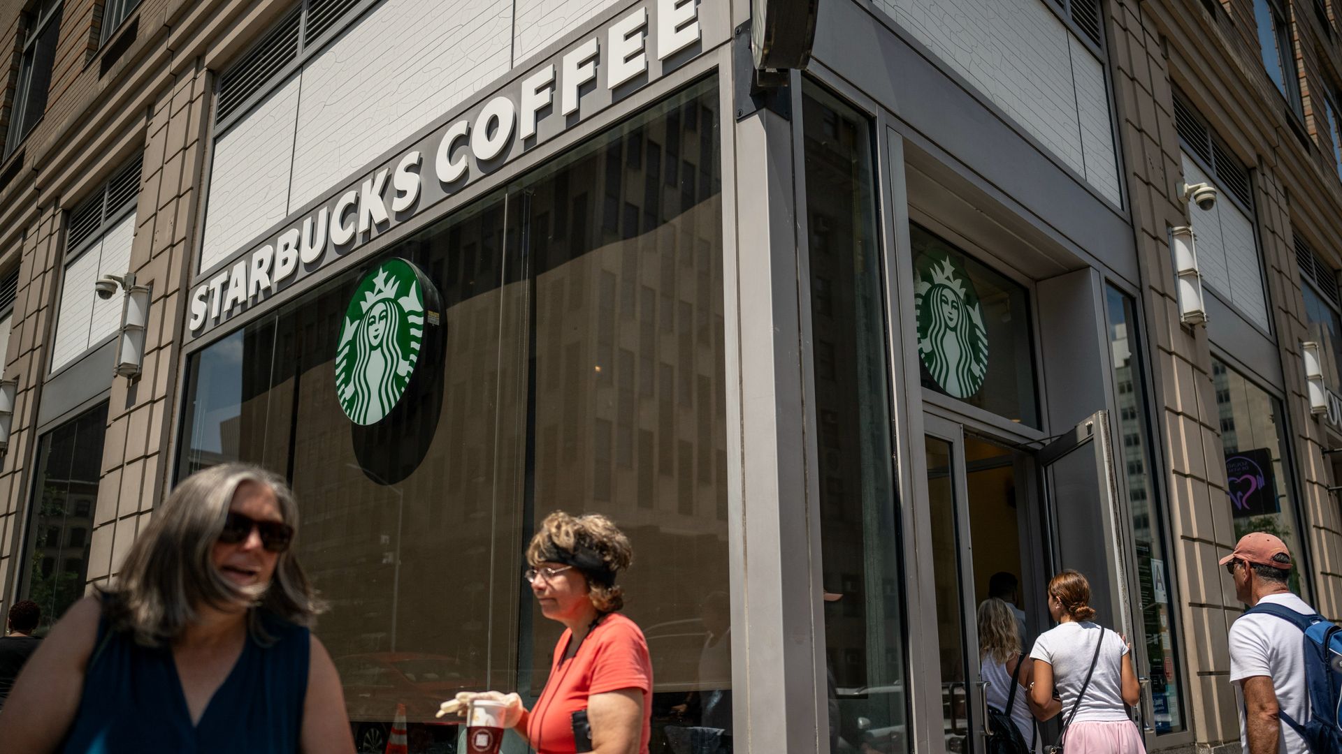 People walk by and into a Starbucks Coffee location. One person that is walking outside has a cup of coffee