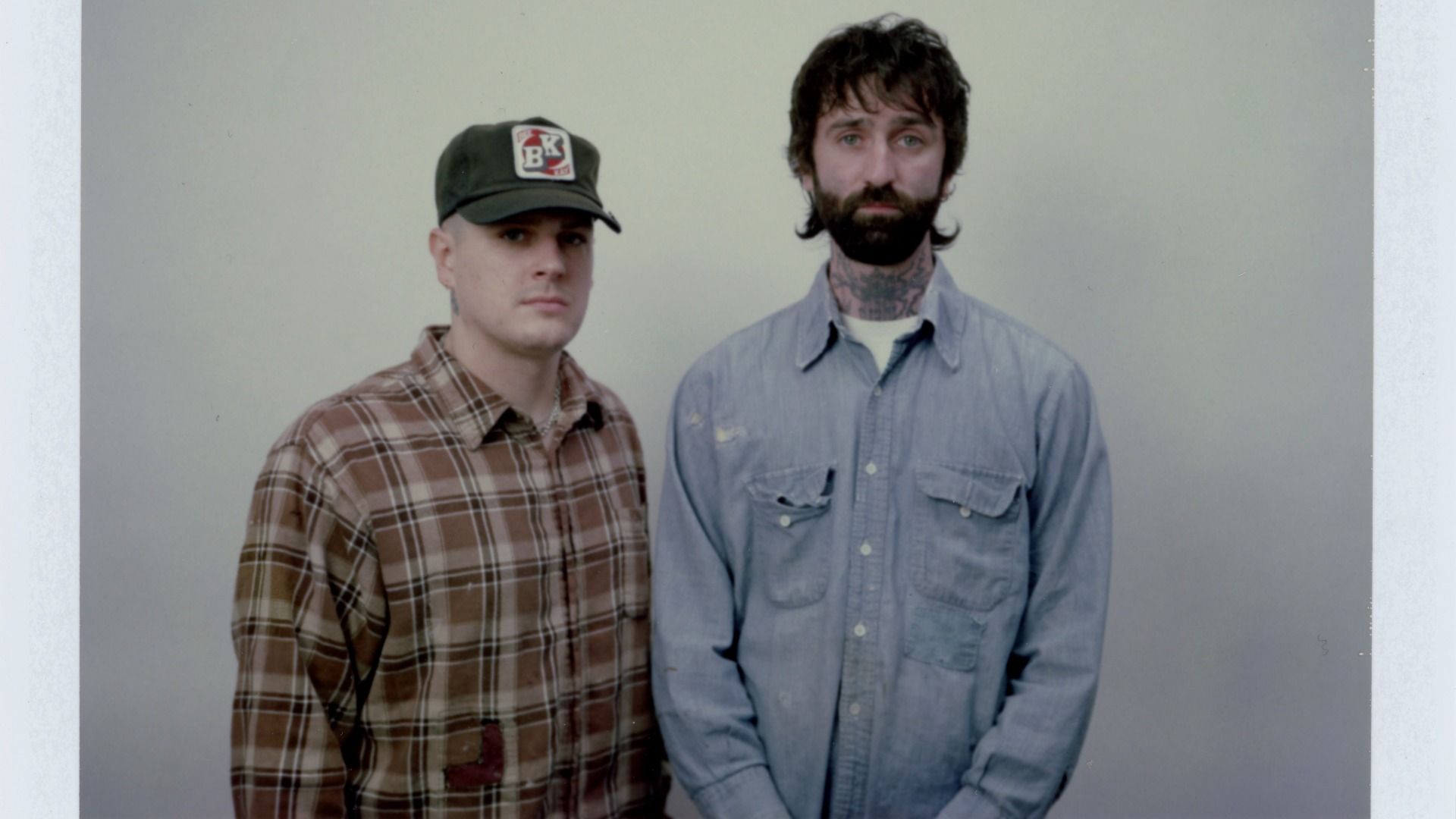 Two men stand side by side against a plain gray wall. Left: brown plaid shirt and green cap with a BK patch. Right: dark beard, neck tattoos, light blue button-down shirt.