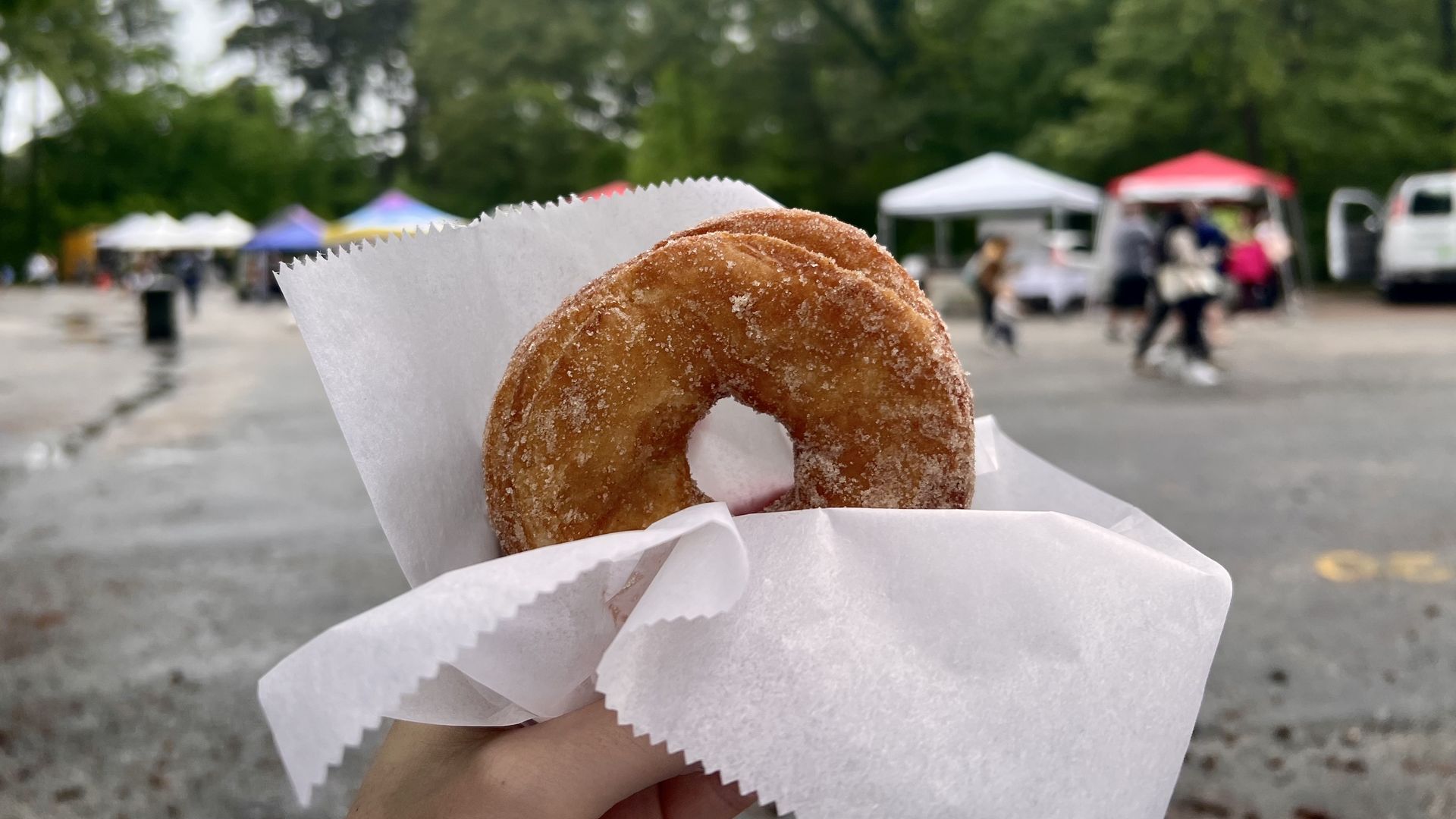A picture of a hand holding up a cronut with a white tissue-like thing and the background, which is a farmers market, is blurred.
