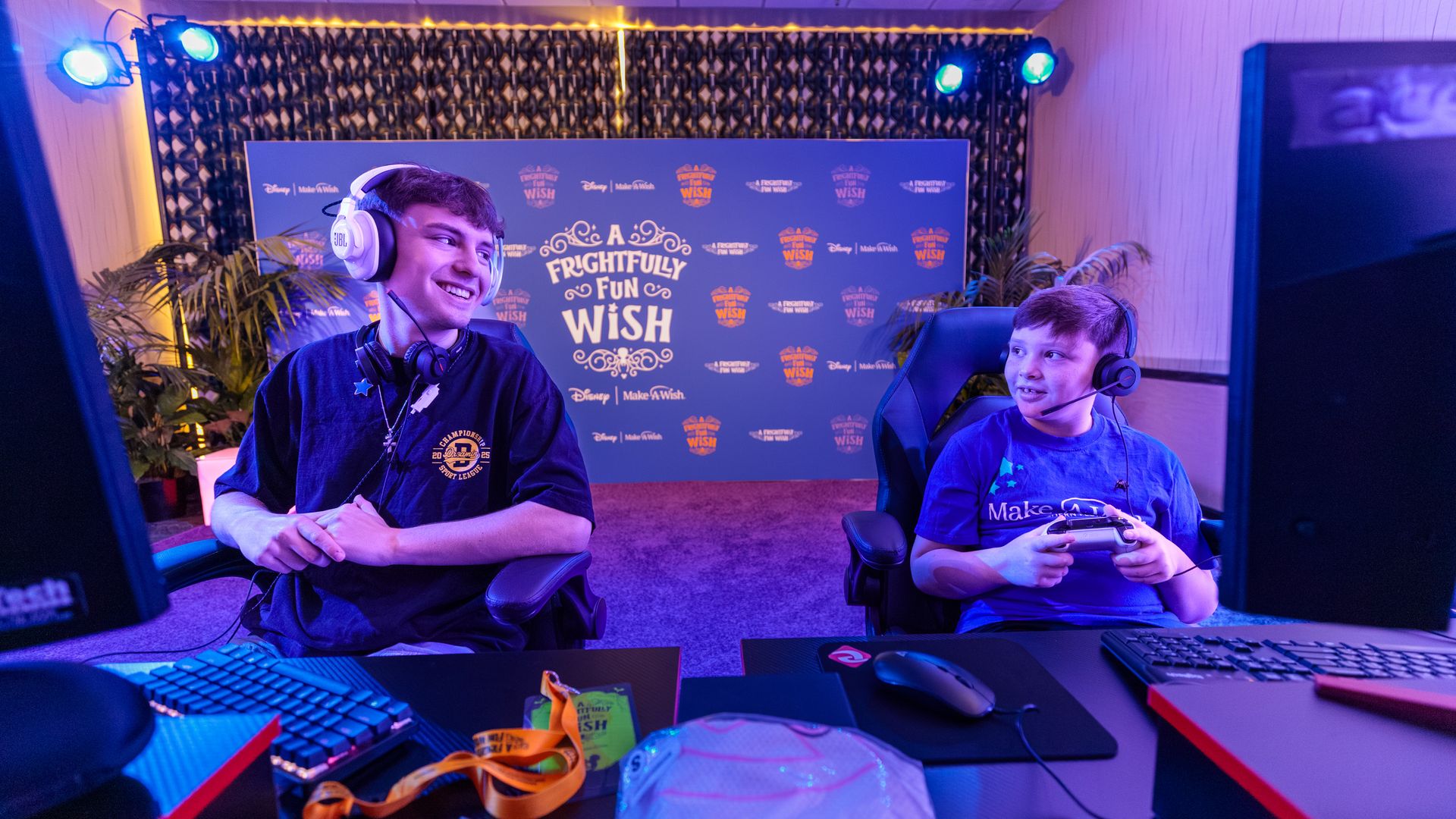 Two boys wearing headsets gaming together in a room with purple lighting and a backdrop reading "A Frightfully Fun Wish" from Disney and Make-A-Wish.