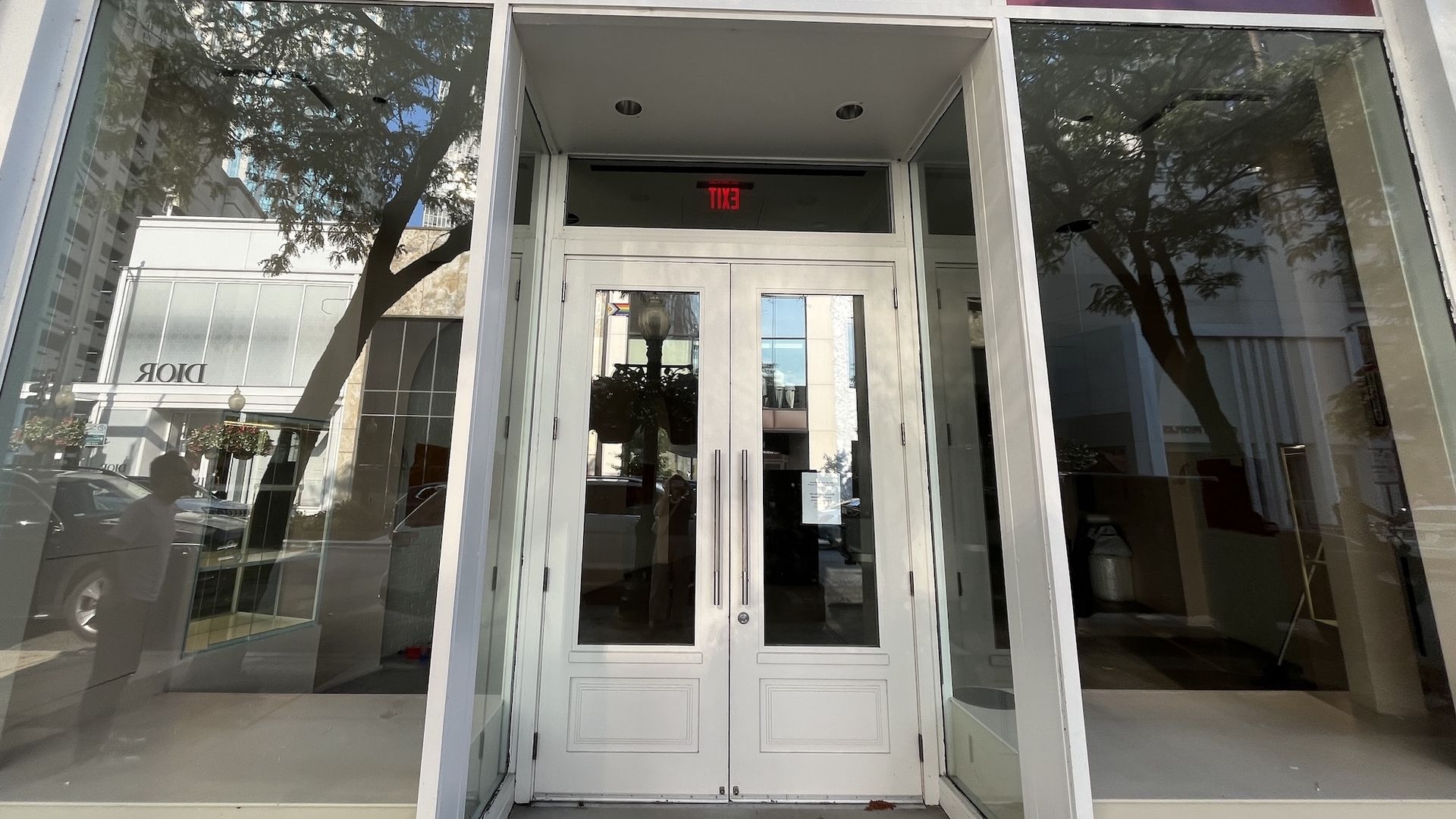 Glass storefront with white framing and double doors; a red EXIT sign above. Reflective glass shows trees and street, with blue and purple banner panels above the entrance.