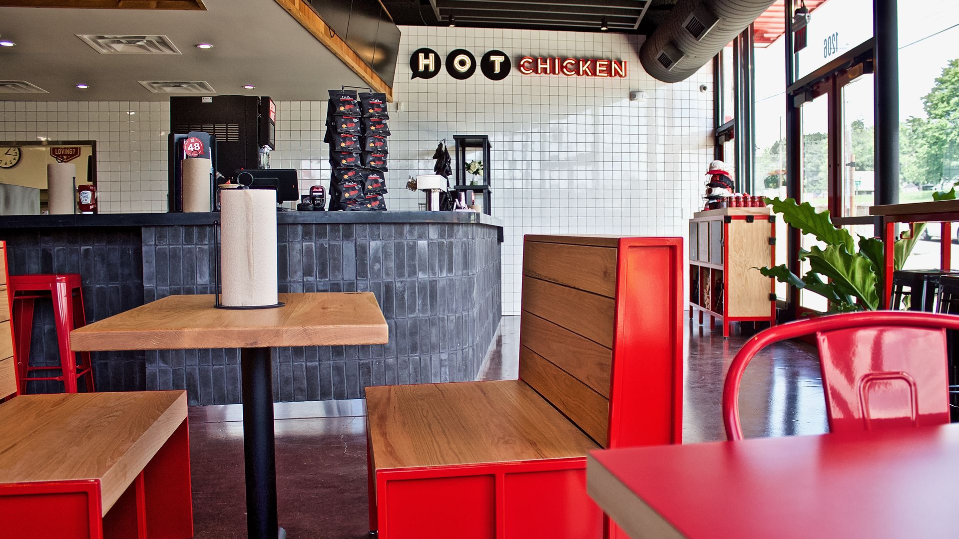 Interior of a fast-casual restaurant with white tile walls, red furniture, and a neon "HOT CHICKEN" sign on the tiled wall; counter area and large windows to the outside.