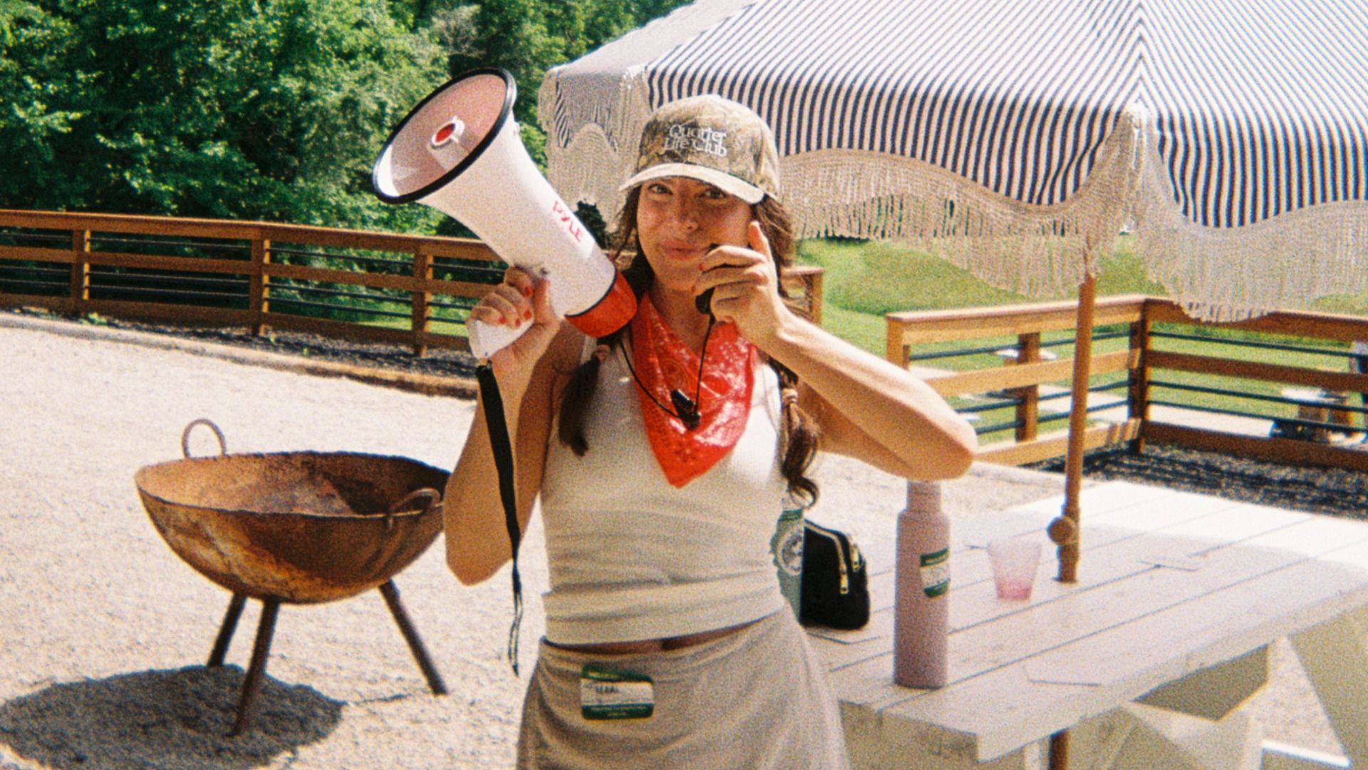 Smiling woman stands outside under striped canopy, holding a white megaphone to her mouth. She wears a camo cap, red bandana, white tank top, and shorts, with a wooden railing and a grill nearby.