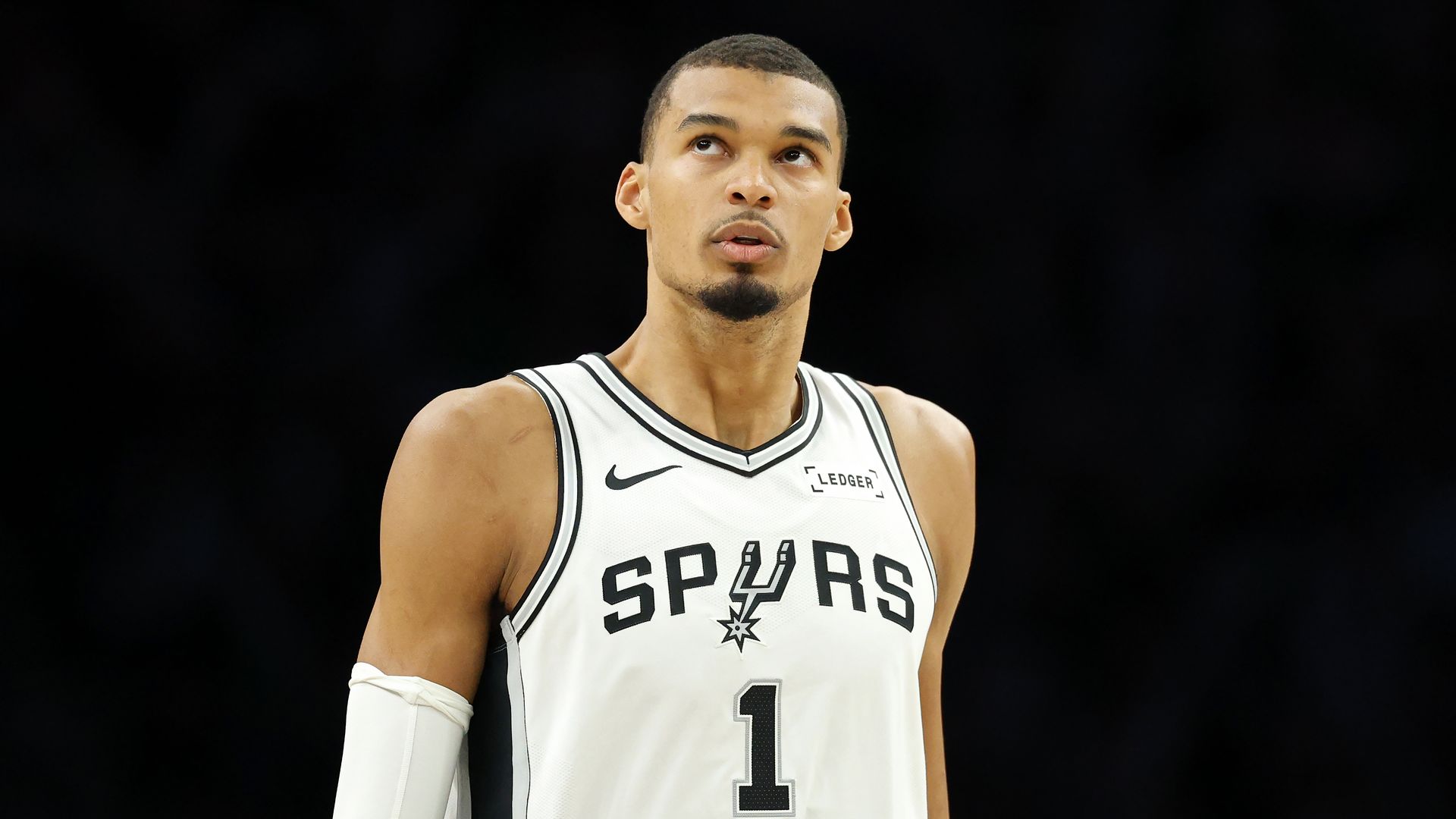 Close-up of a male basketball player in a white San Antonio Spurs jersey (number 1) with black trim, looking upward, against a dark arena background.