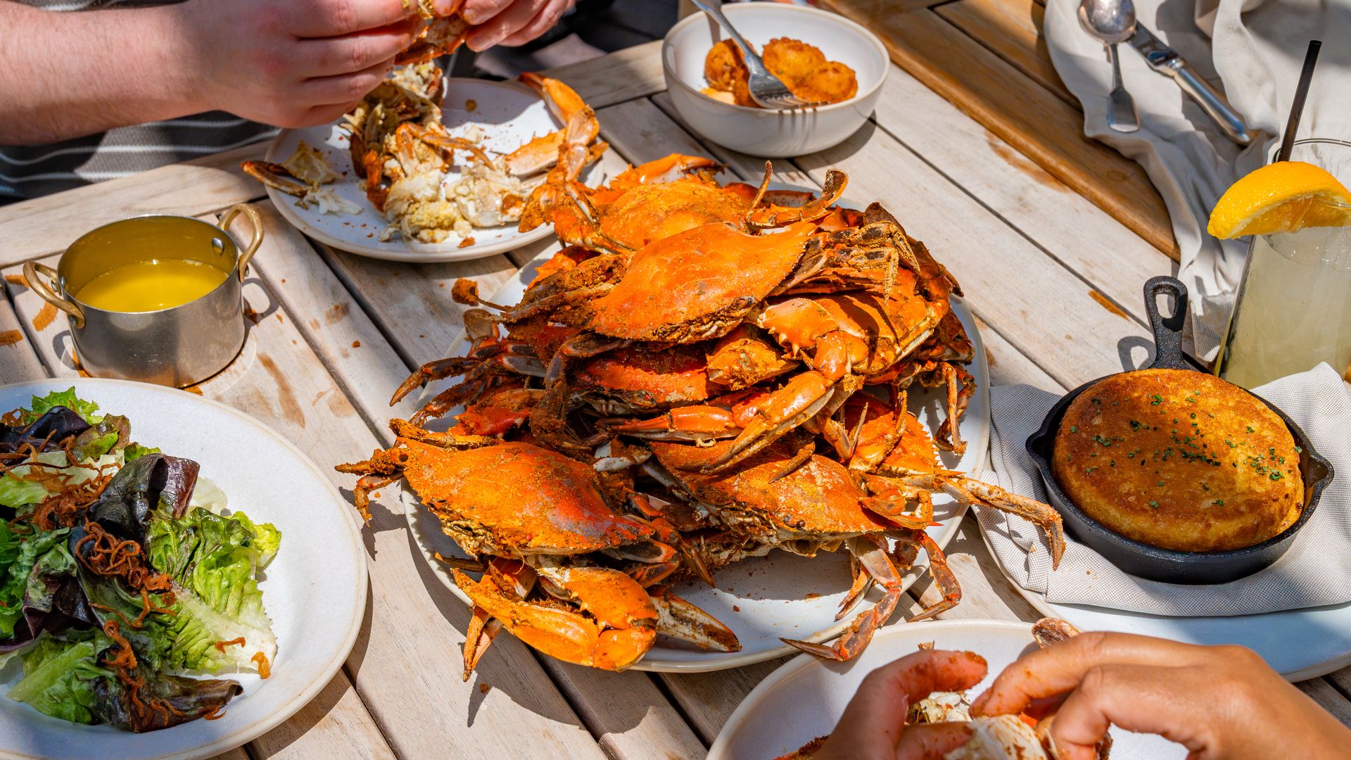 Outdoor wood table seafood feast: bright orange crabs heaped on plates, bowls of crab meat and corn, a lemon drink, greens, and hands cracking shells.