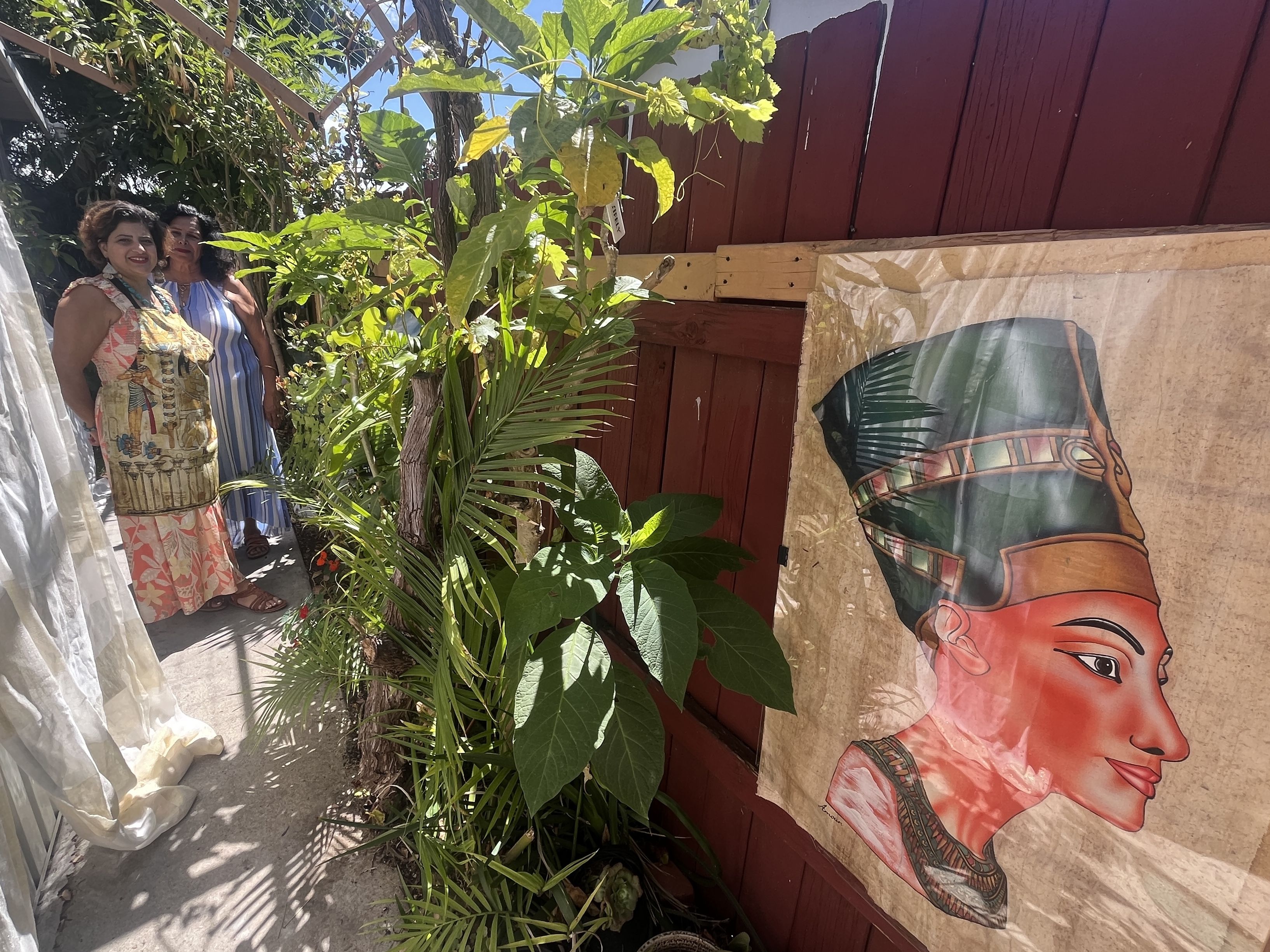 Two women stand beside a maroon wooden fence, surrounded by lush plants. One wears a colorful floral dress, the other a blue striped dress. A large poster showing a car and a woman's face hangs on the fence.