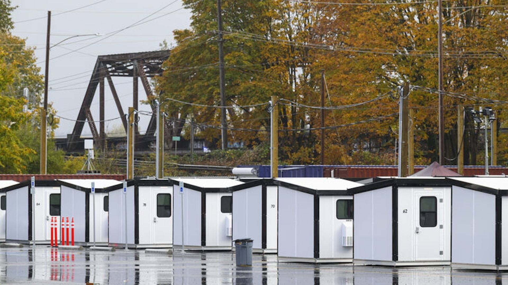 A photo of tiny home shelters in the foreground, with a steel bridge and fall foliage trees in the background.