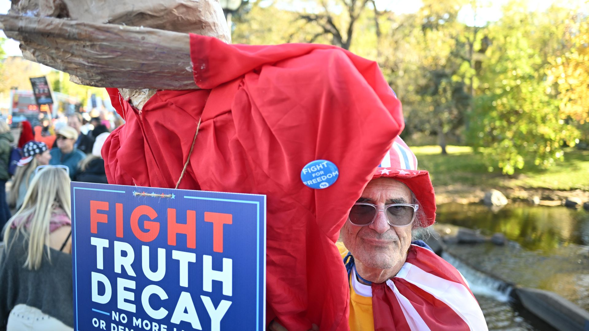 Man wearing an American flag-themed hat and cape stands by a sign saying "Fight Truth Decay" in red, white, and blue, with a crowd and green trees in background.