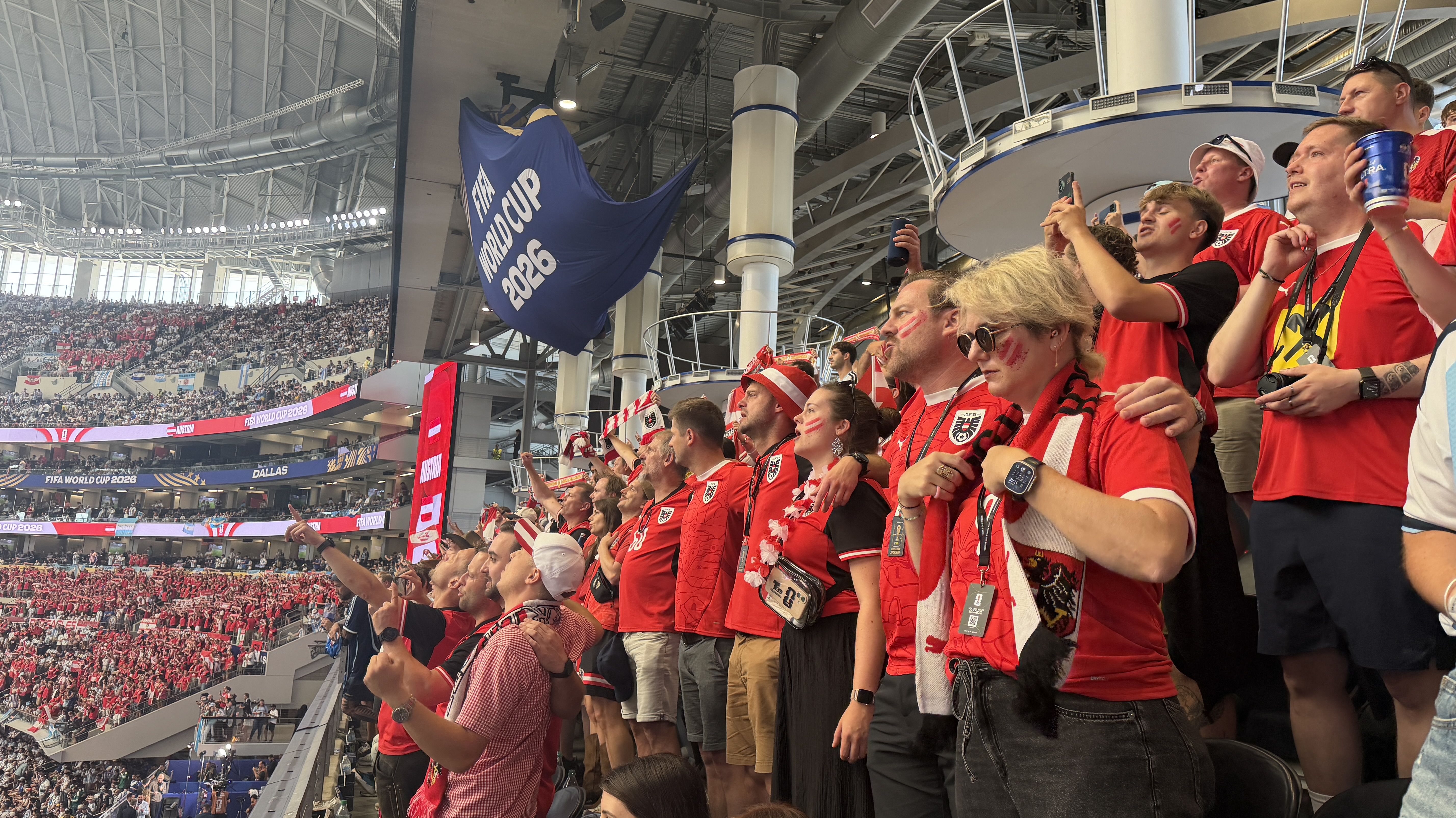 Crowd of red-clad soccer fans in a large stadium, many filming with phones and cheering. A large blue FIFA World Cup 2026 banner hangs from the upper level amid steel beams.