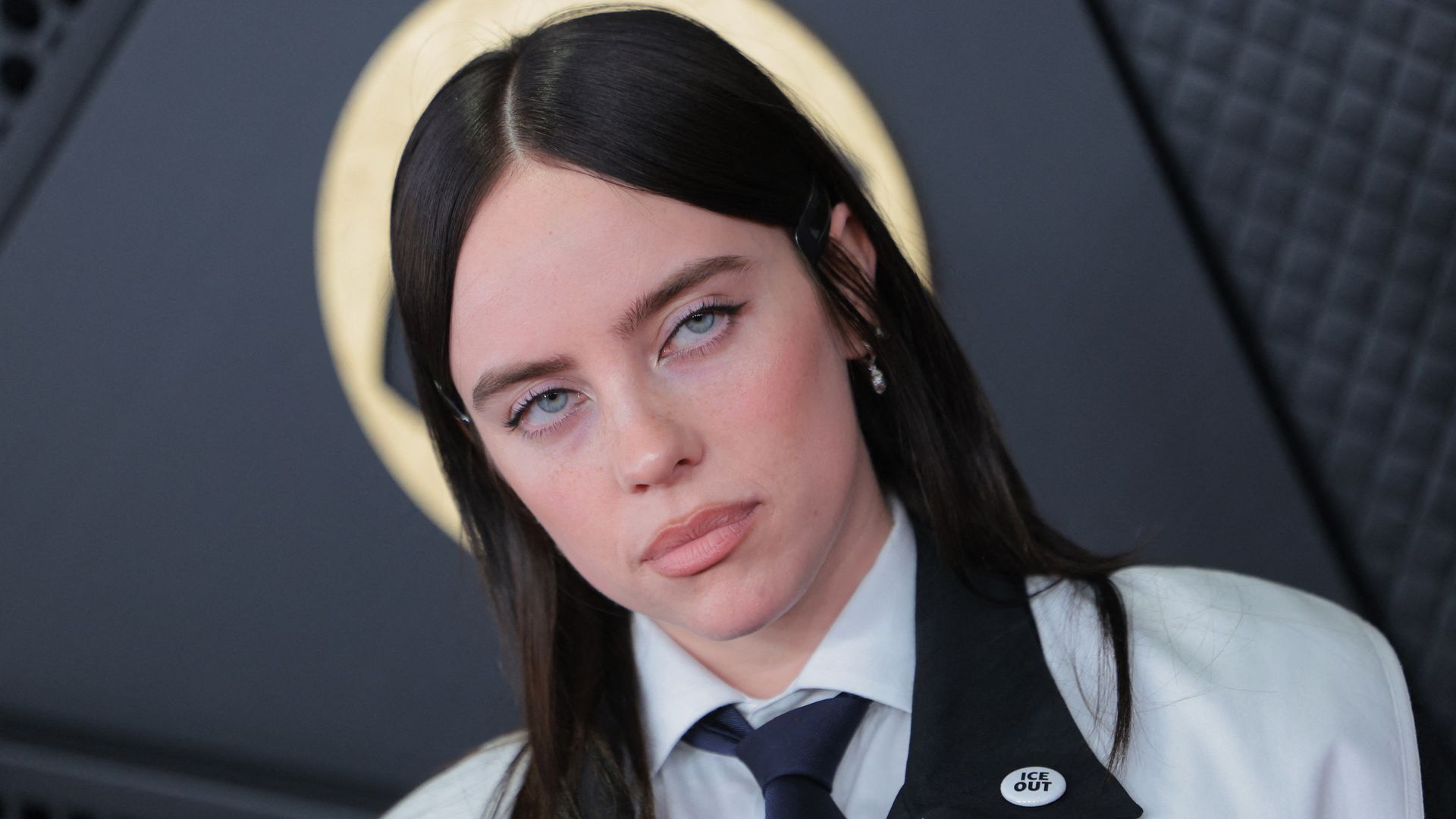 Close-up of a woman, singer songwriter Billie Eilish, with blue eyes, brown hair, wearing a white shirt, dark tie, and jacket with an "ICE OUT" pin, against a dark blurred background with a gold circular shape.