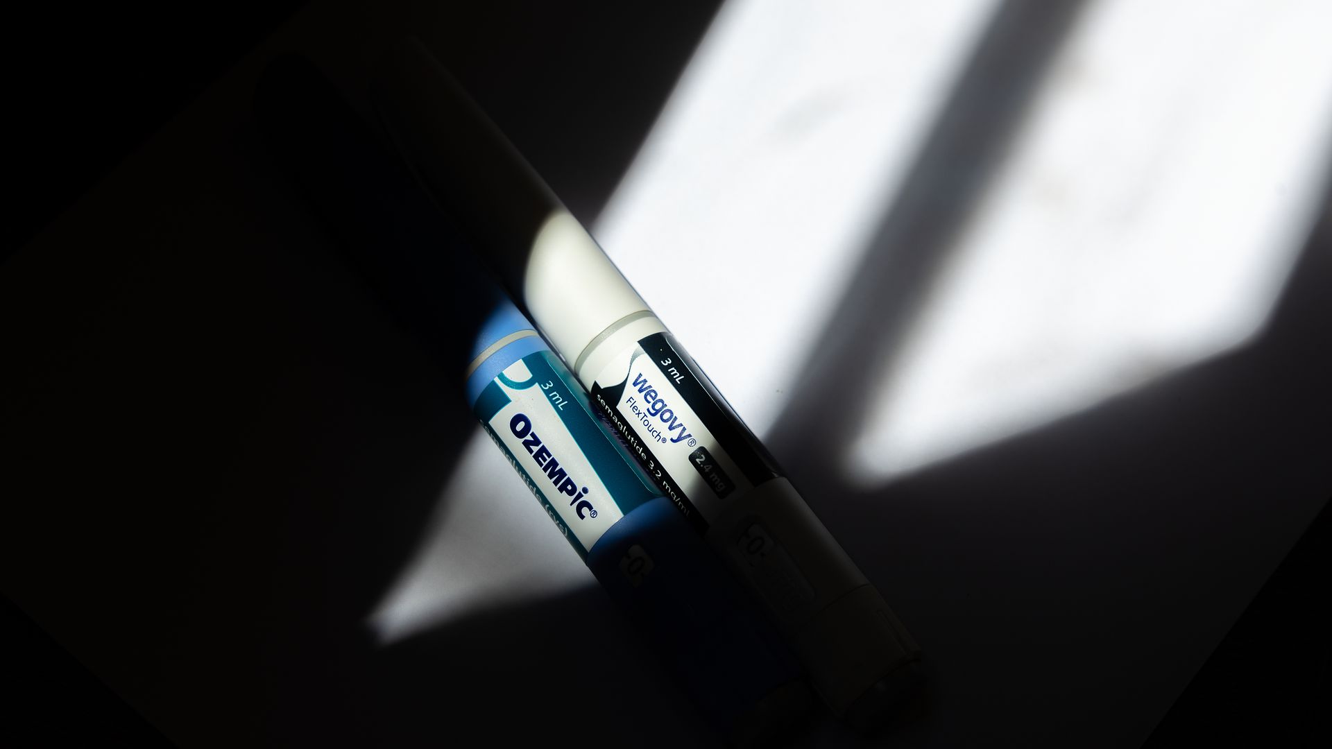 Two medication pens labeled Ozempic and Wegovy in blue and white, lying on a dark surface with a bright shadow pattern across them.