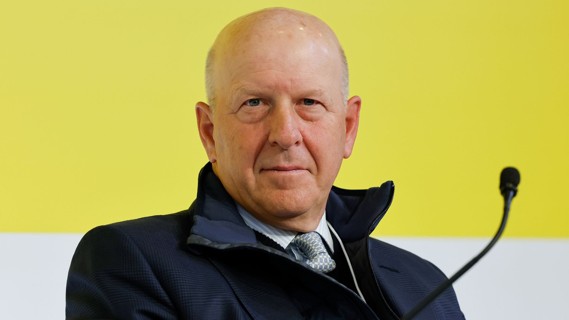 bald man wearing a jacket over a suit and tie with a yellow and white background behind him