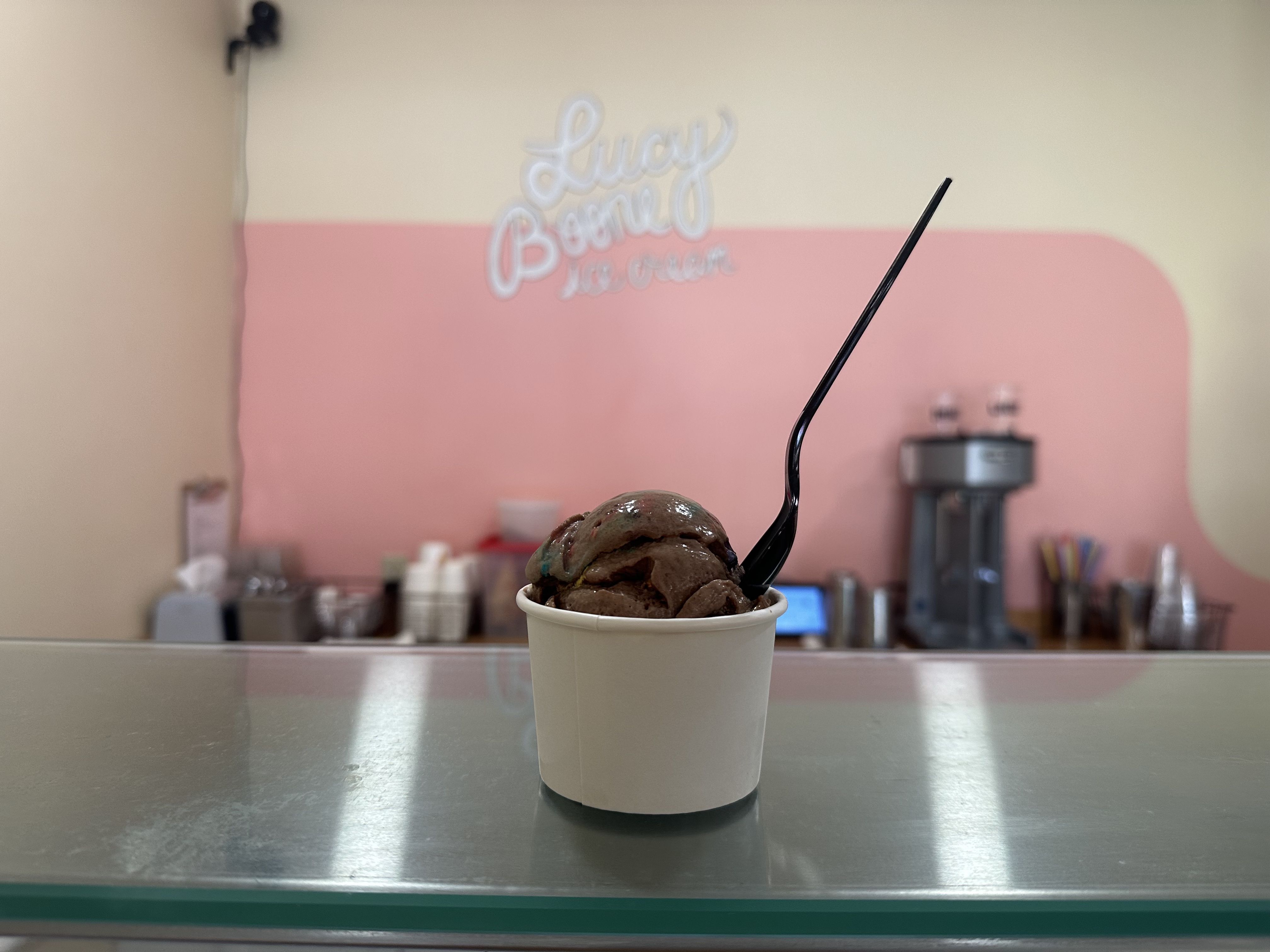 A scoop of cosmic brownie ice cream sits on the counter at Lucy Boone. The shop's logo is painted on the wall behind the cup of ice cream, which has a black plastic spoon sticking out of it. 