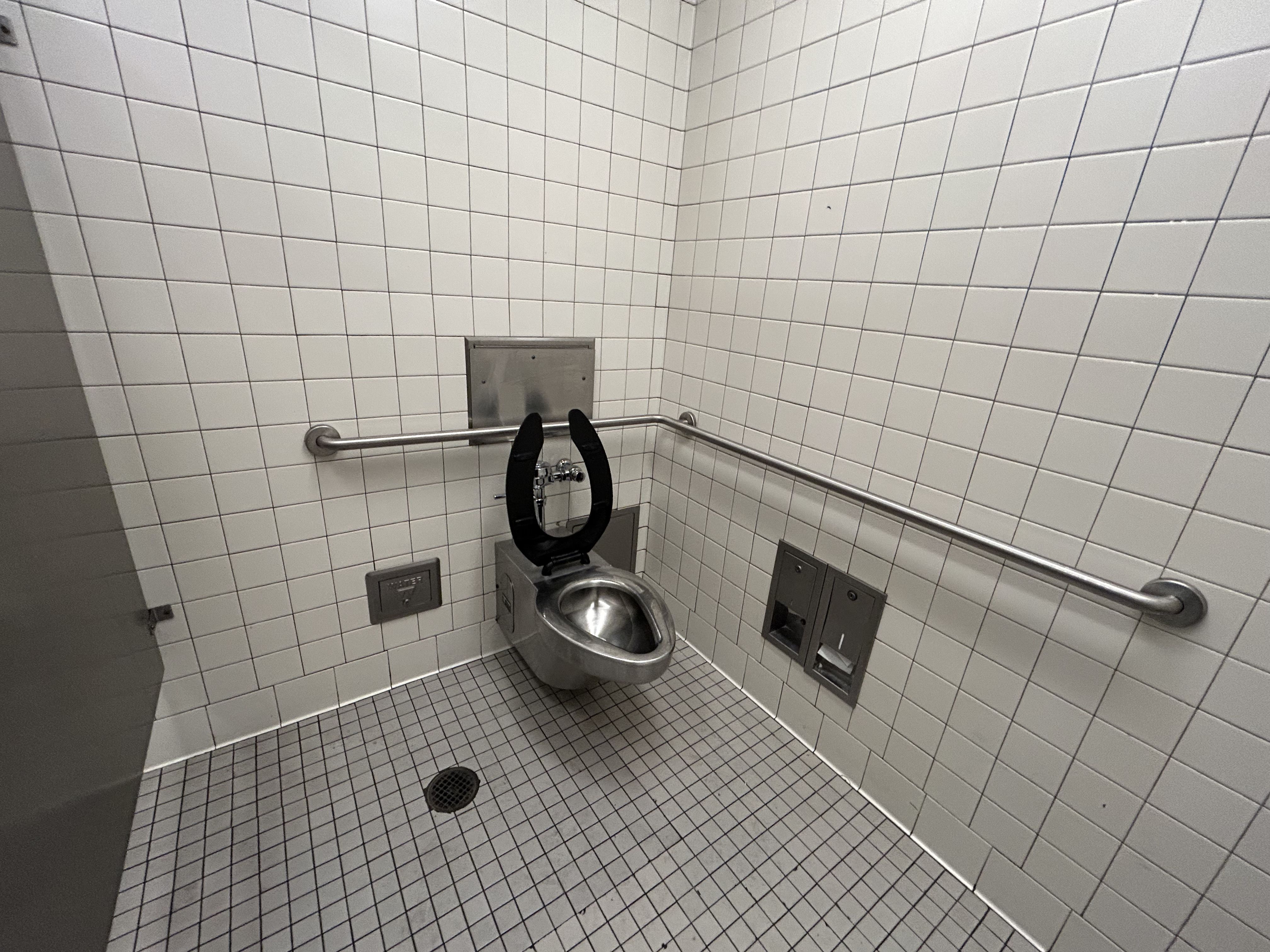 Accessible public restroom stall with white tiled walls and gray floor. Stainless toilet, black seat, lid open. Parallel grab bars along walls, flush plate, floor drain, and a paper dispenser nearby.