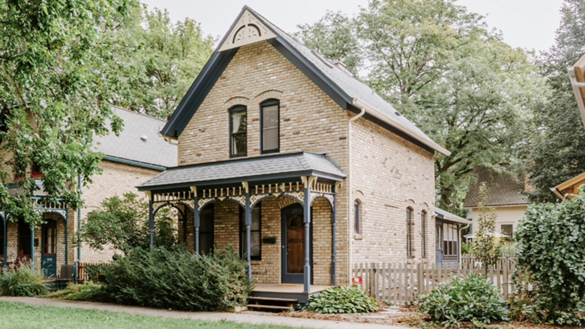 Two-story beige brick house with dark blue trim, a covered front porch, surrounded by green bushes and trees on a grassy lawn under a cloudy sky.