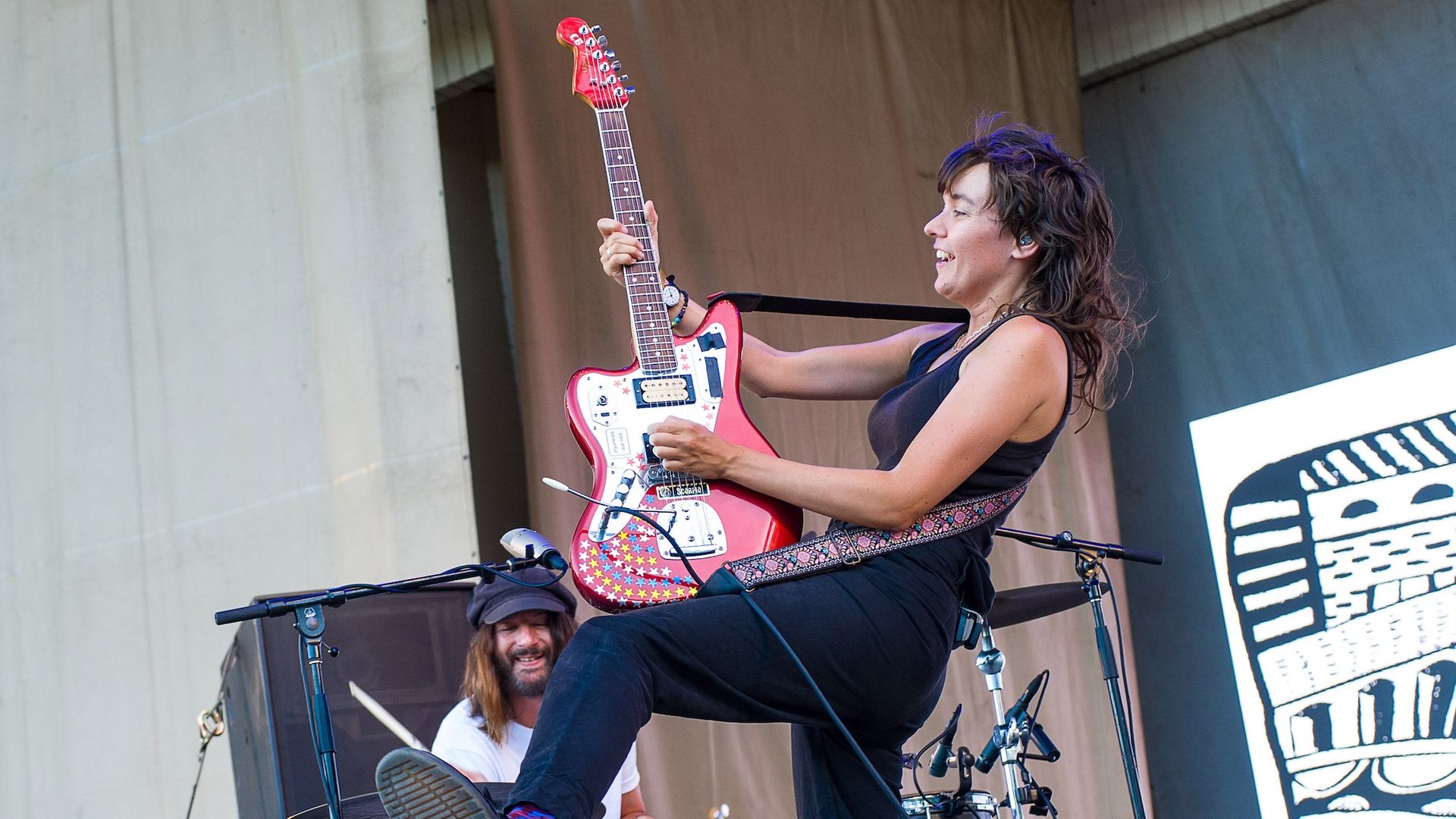 Courtney Barnett wearing all black and playing a red and white guitar while a drummer plays in the background.