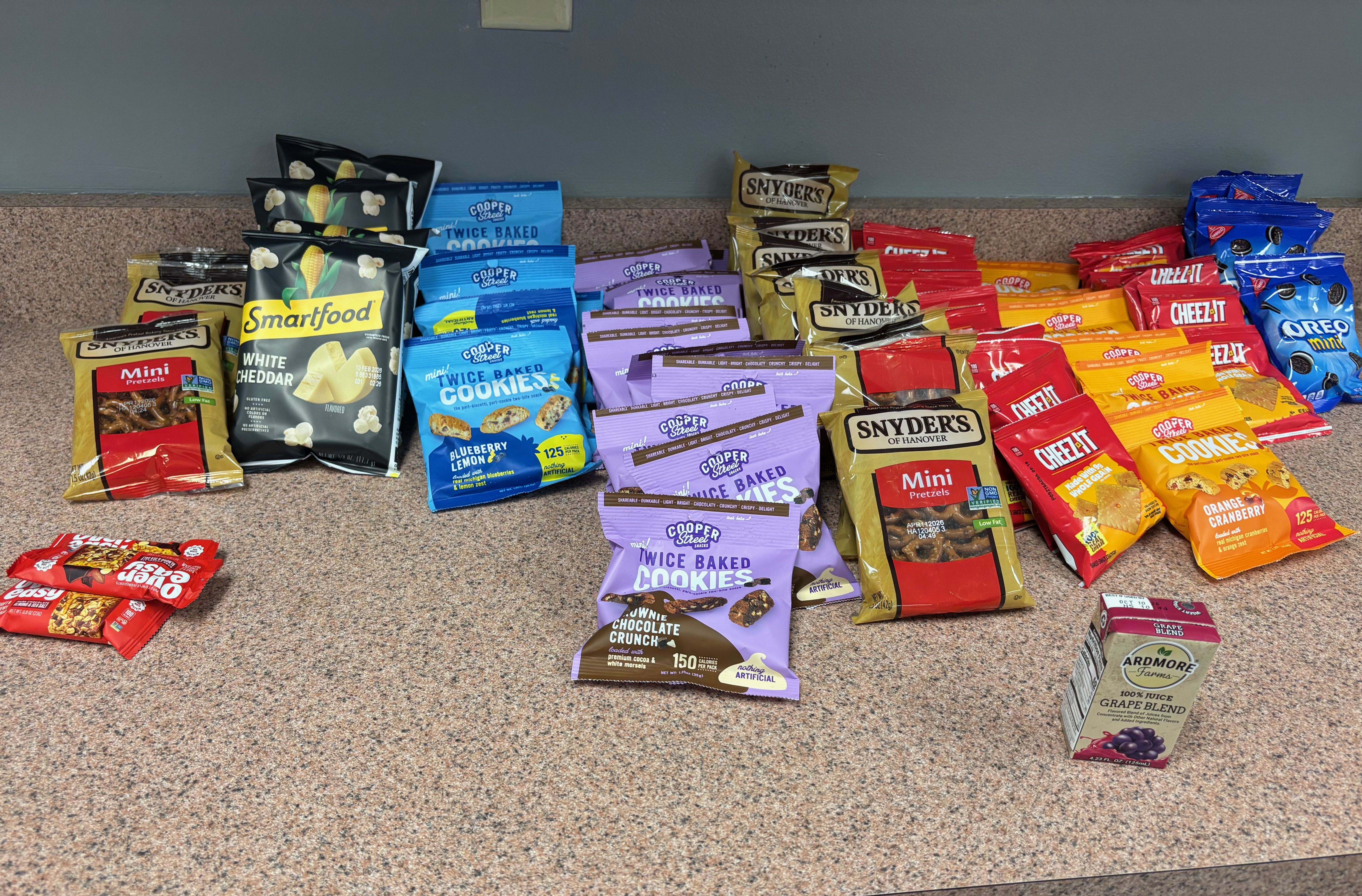 Assorted snacks on a countertop including Smartfood white cheddar popcorn in black bags, Cooper Street Twice Baked Cookies in blue and purple, Snyder's Mini Pretzels in yellow, Cheez-It crackers in red, and Oreo Mini in blue packs, with a small Ardmore Farms grape juice box.