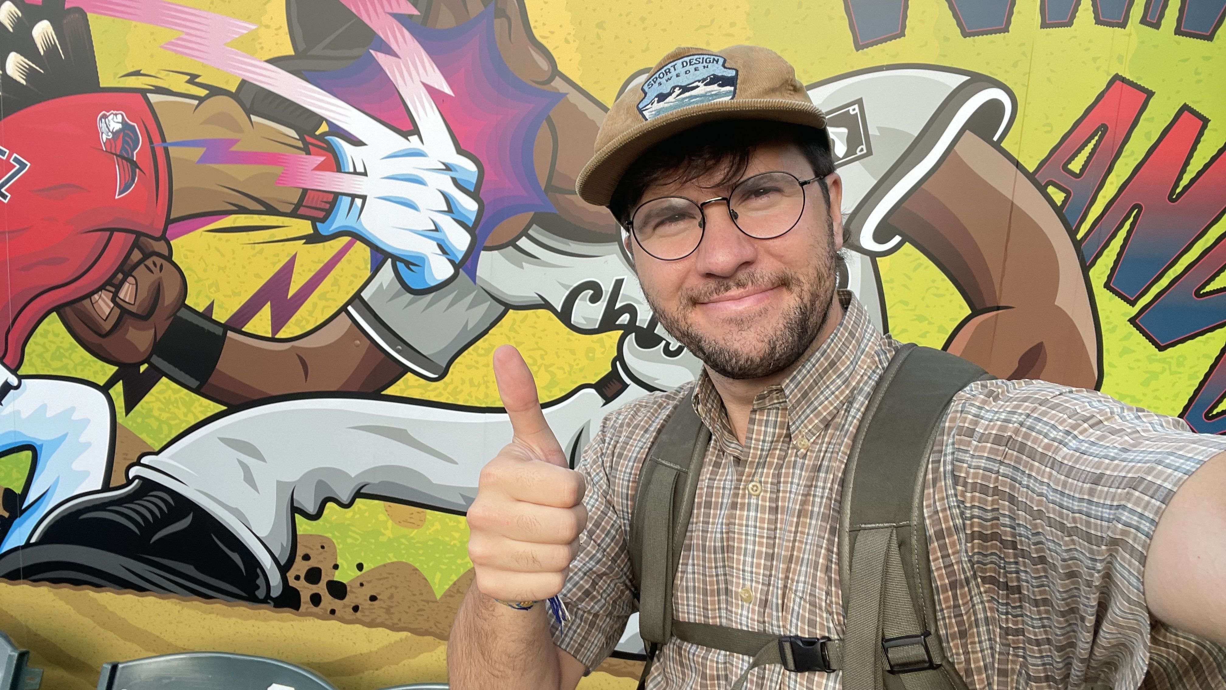 Selfie of a man with round glasses and a plaid shirt, wearing a brown cap with a "Sport Design" patch, and backpack straps, giving a thumbs-up in front of a colorful graffiti mural with a boxing glove and cartoon figures.