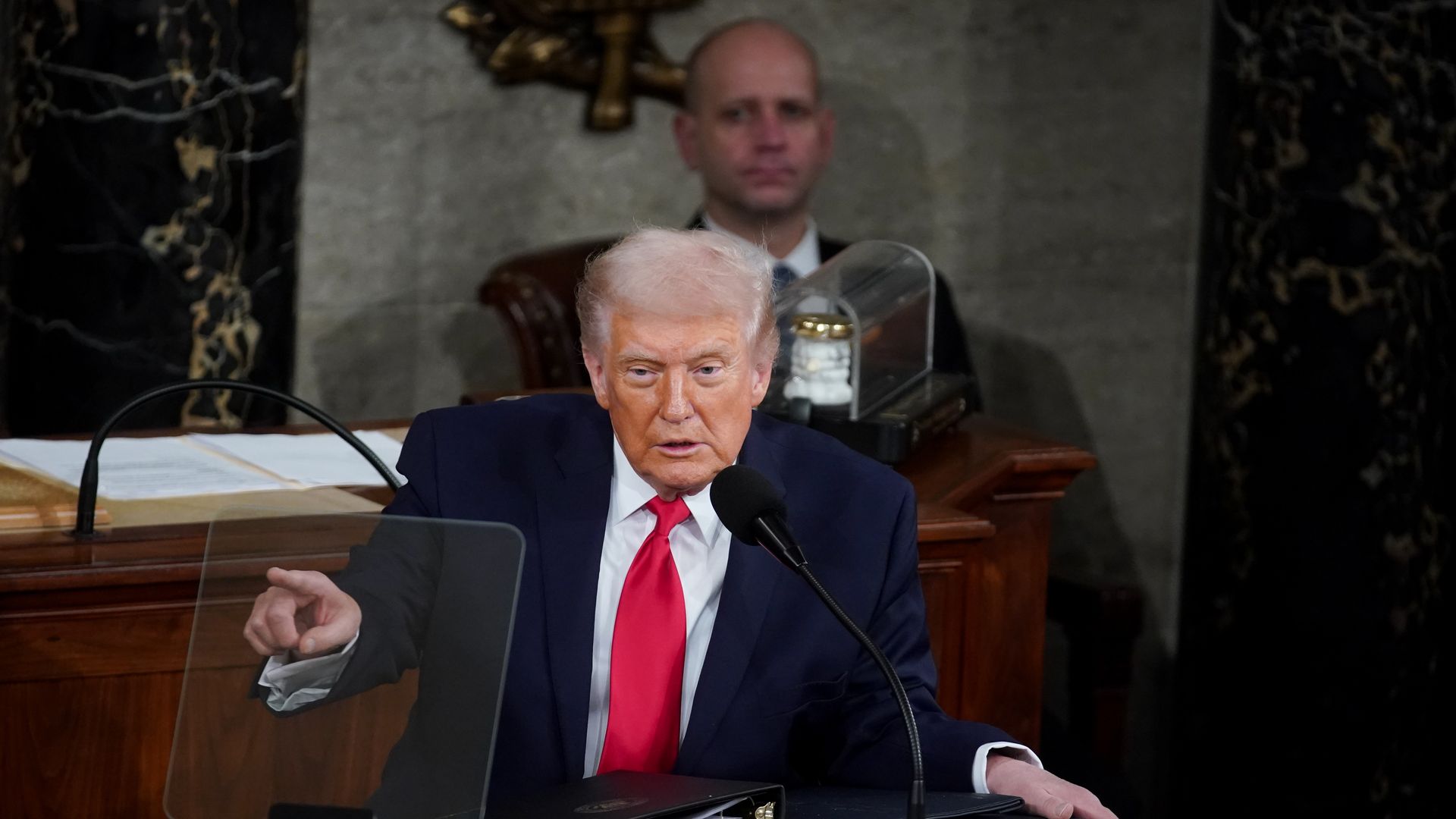 President Trump, with white hair in a dark suit and red tie, speaks at a wooden podium with microphone and a clear teleprompter, pointing with his left hand; a man sits blurred in the background.
