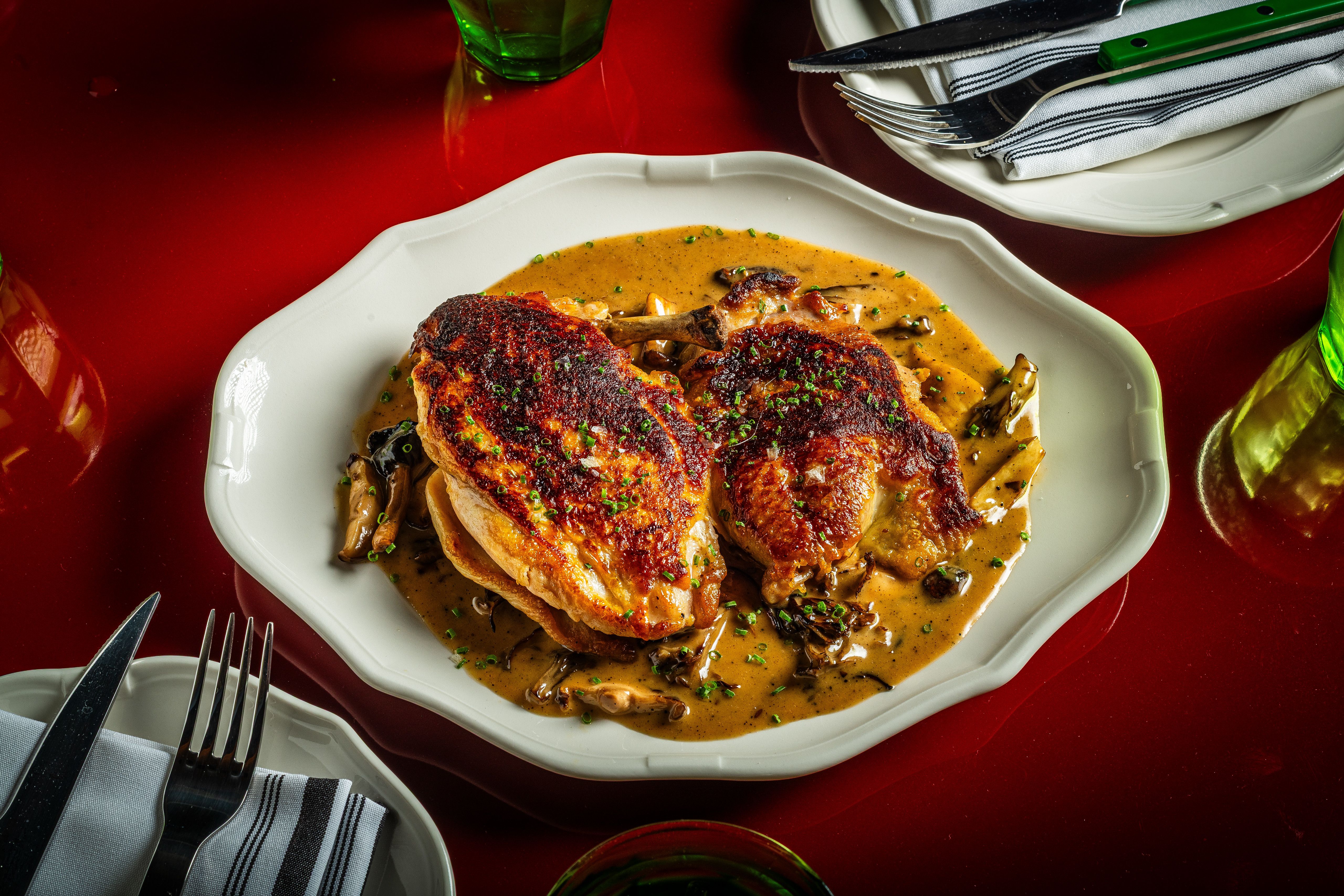 White plate with crispy roasted chicken in creamy mushroom sauce, garnished with chopped green herbs, on a red table; forks, knife, napkin, and a green glass nearby.