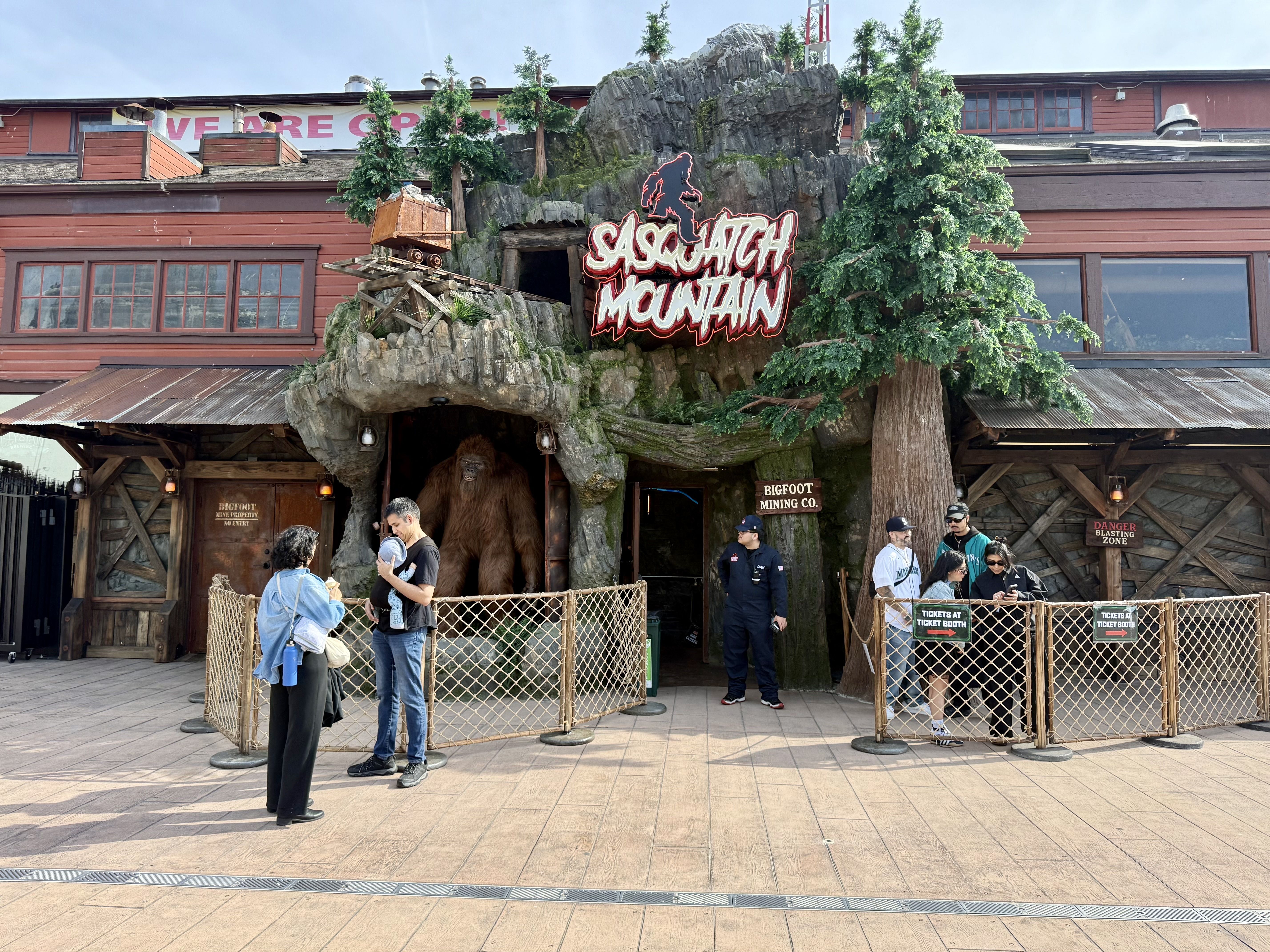 Entrance to "Sasquatch Mountain" ride: a rocky mountain facade with a neon red Sasquatch sign, wooden mining exterior, a tilted mine cart on tracks, and visitors behind a fencing barrier.