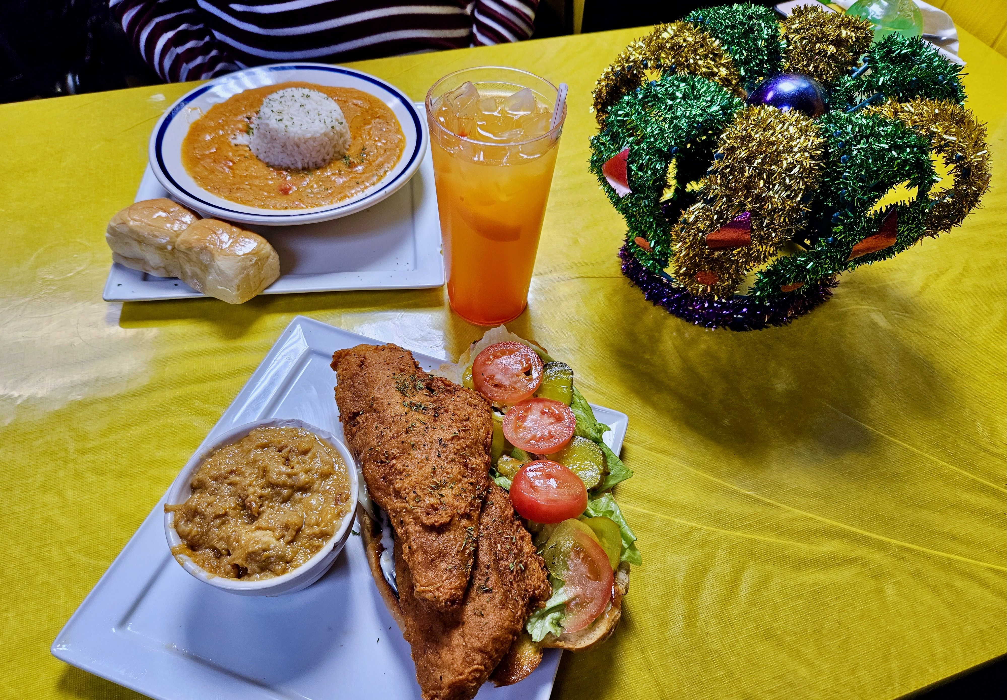 Two white plates (with a po'boy and crawfish étoufée) on a yellow tablecloth next to a Mardis Gras crown decoration and glass of punch