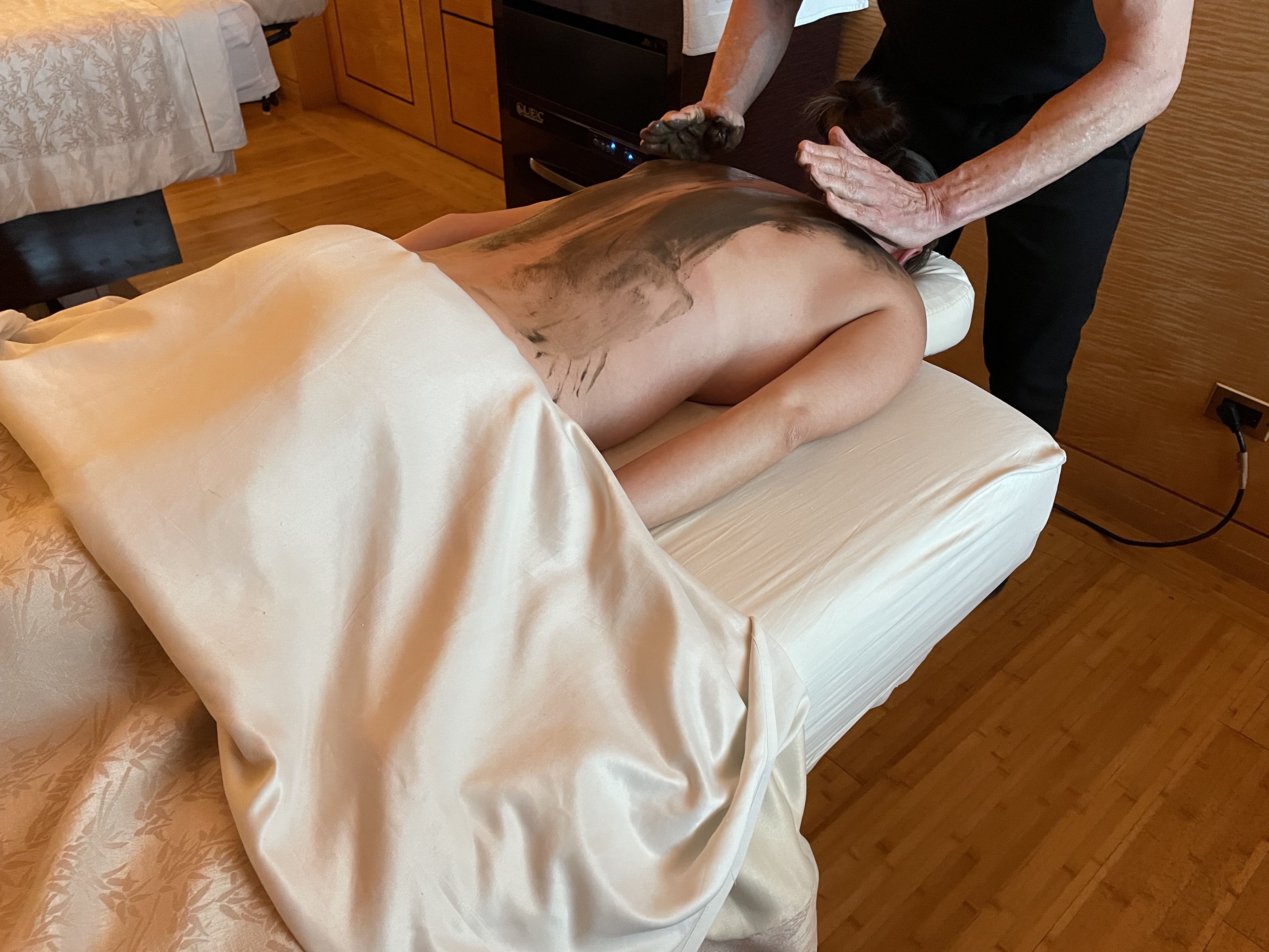 A massage therapist applies a CBD-infused mud mask on the back of a woman during a spa treatment.