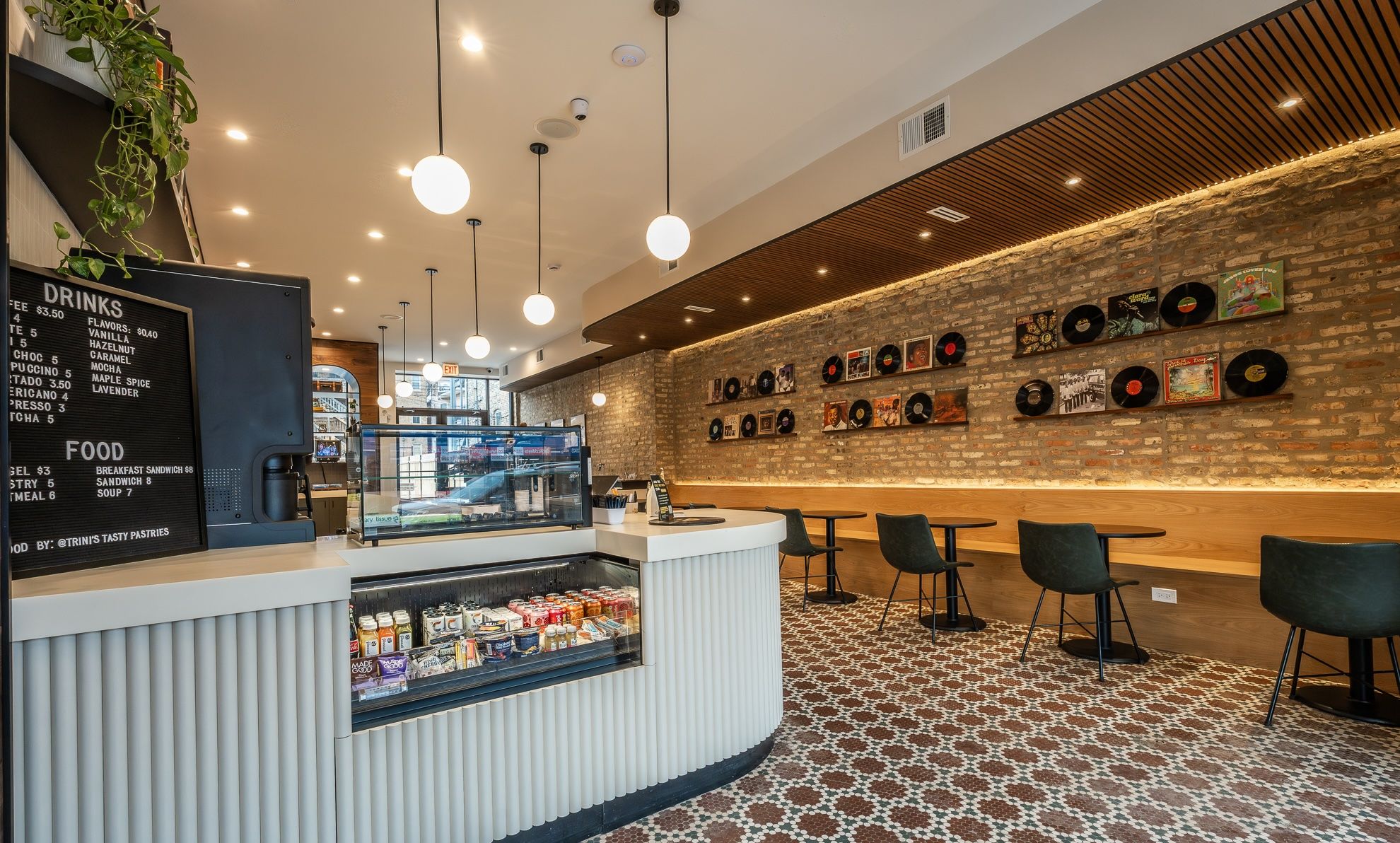 Bright cafe interior featuring a curved white counter with a glass beverage cooler, a brick wall lined with vinyl records, round pendant lights, and black chairs along a wooden banquette.