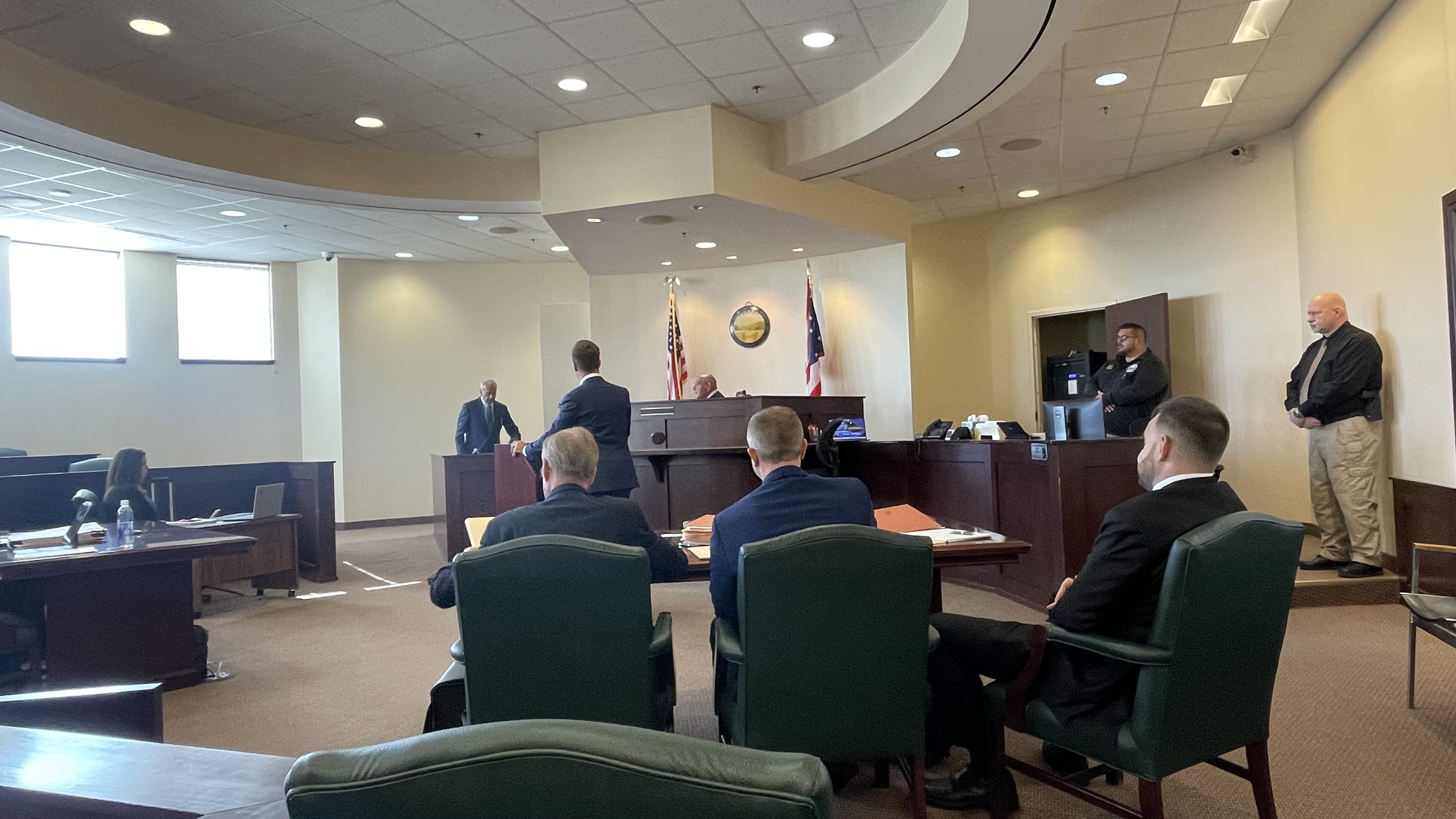 Courtroom scene with a judge's bench and two flags behind it. Suited attendees sit as a man speaks at the lectern; guards stand along the right wall, bright windows on the left.