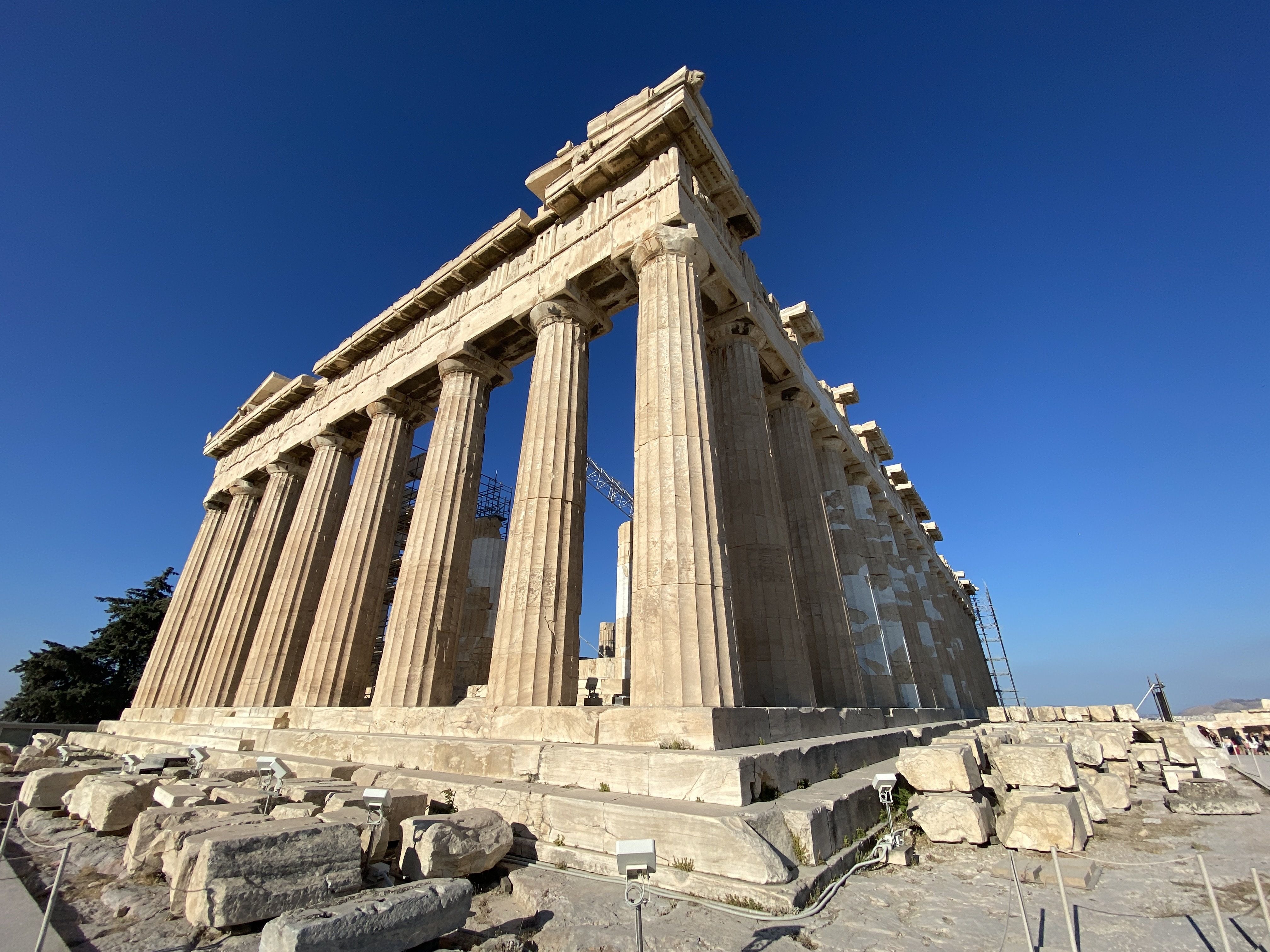 The Parthenon temple in Athens, Greece, shown from a low angle against a clear blue sky, with beige stone columns and restoration scaffolding visible inside.