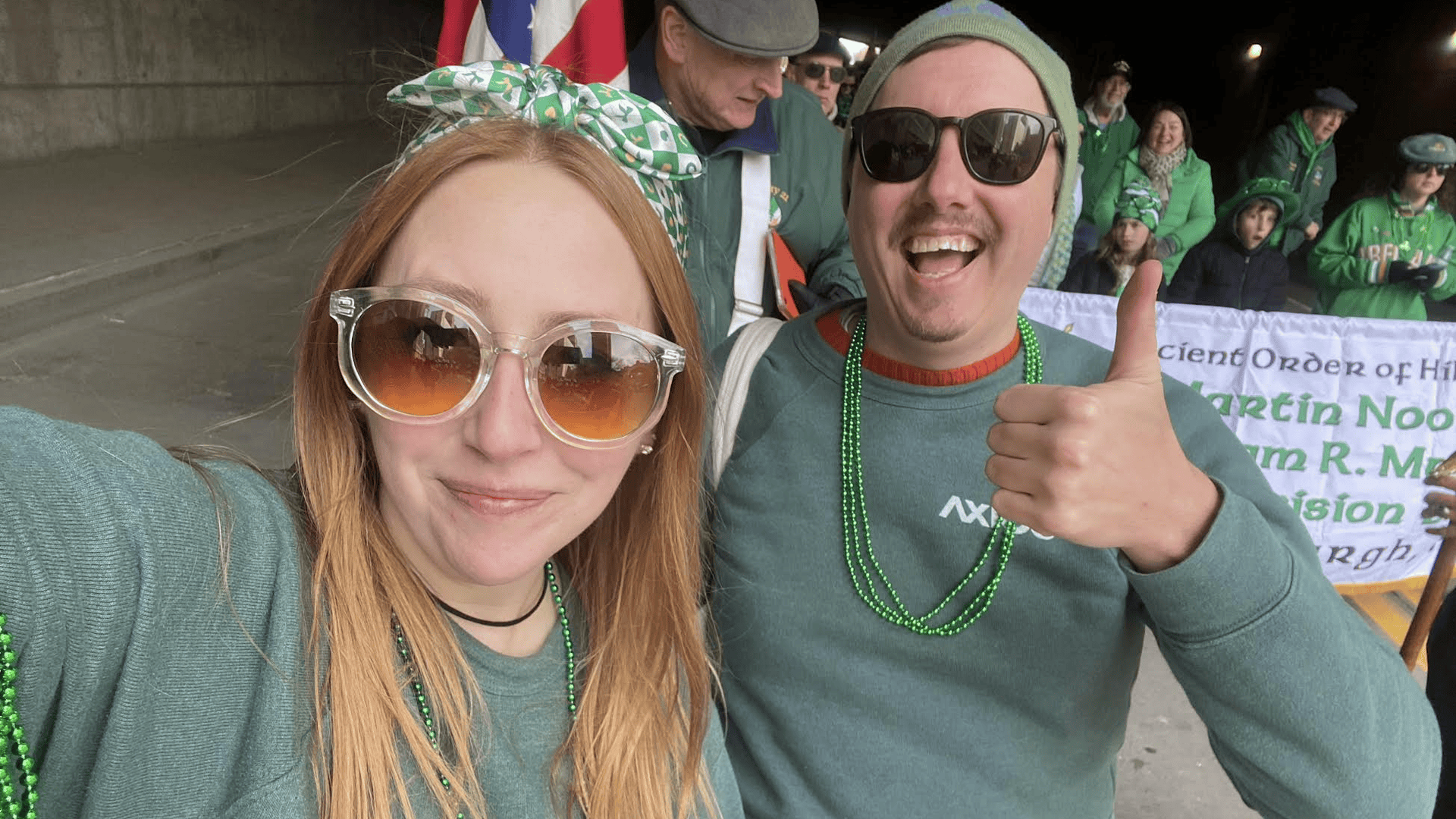 Selfie at a parade: a red-haired woman in oversized sunglasses with a green headband and beads, beside a man in a green beanie and sunglasses giving a thumbs up; crowd in green behind.