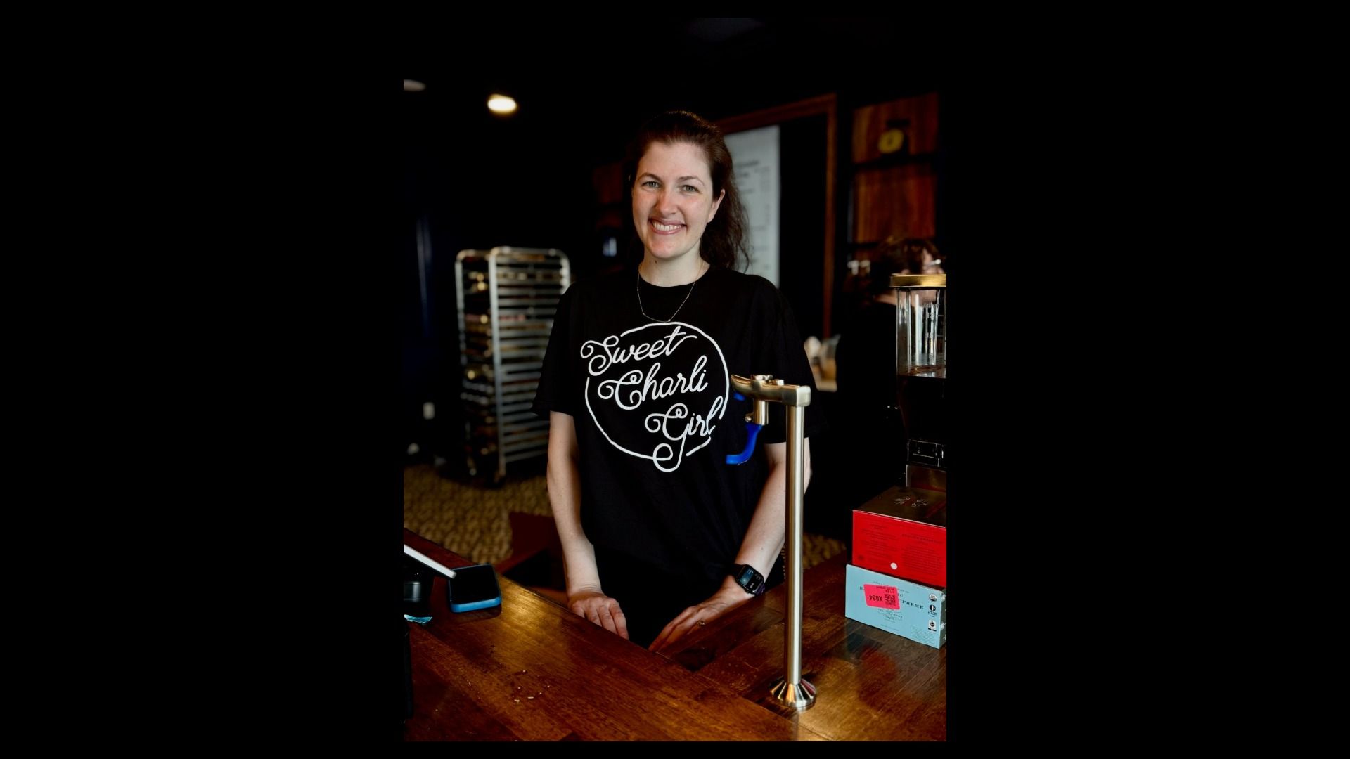 Smiling woman behind a wooden counter in a dim cafe, wearing a black T-shirt that reads "Sweet Charli Girl", with cafe equipment and shelves in the background.