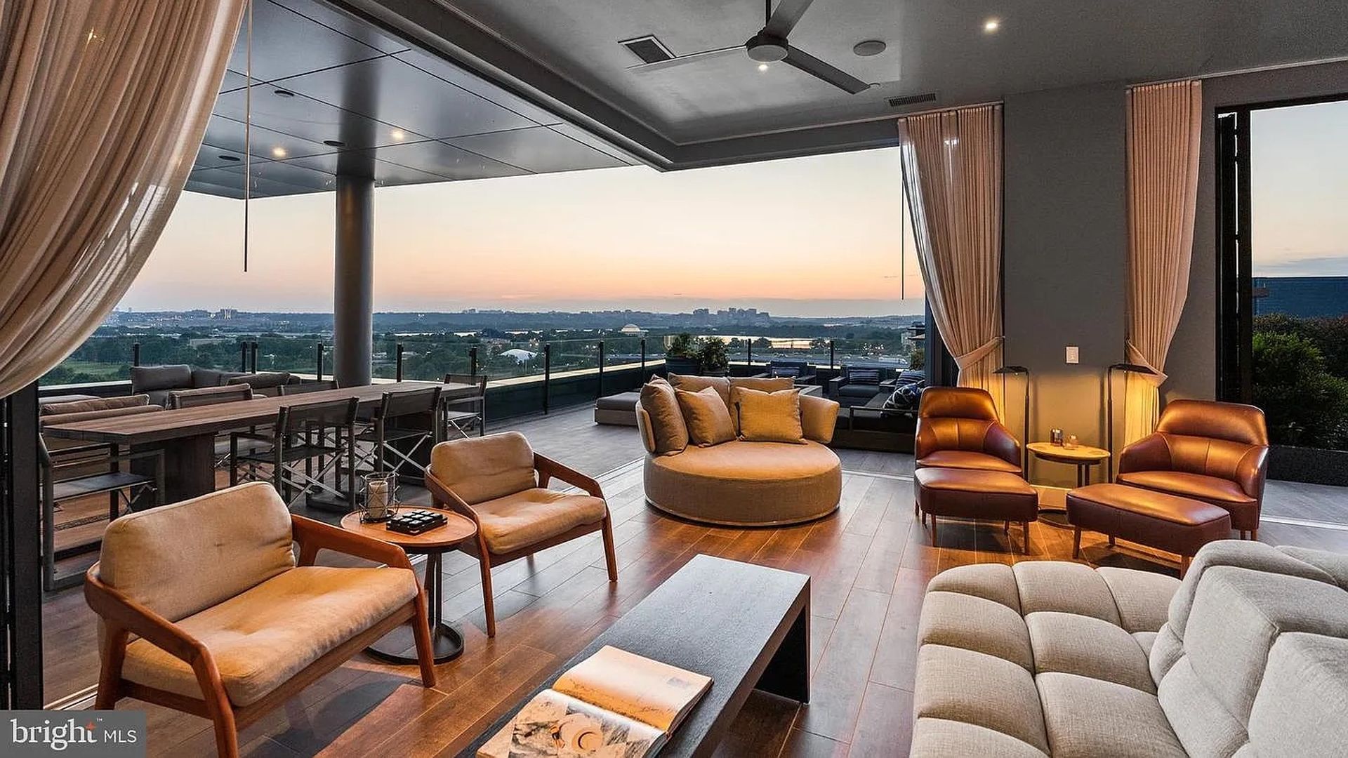 Modern rooftop lounge at sunset with beige sofas, brown lounge chairs, a circular ottoman, a long dining table, sheer curtains, and glass railing overlooking a distant city skyline.