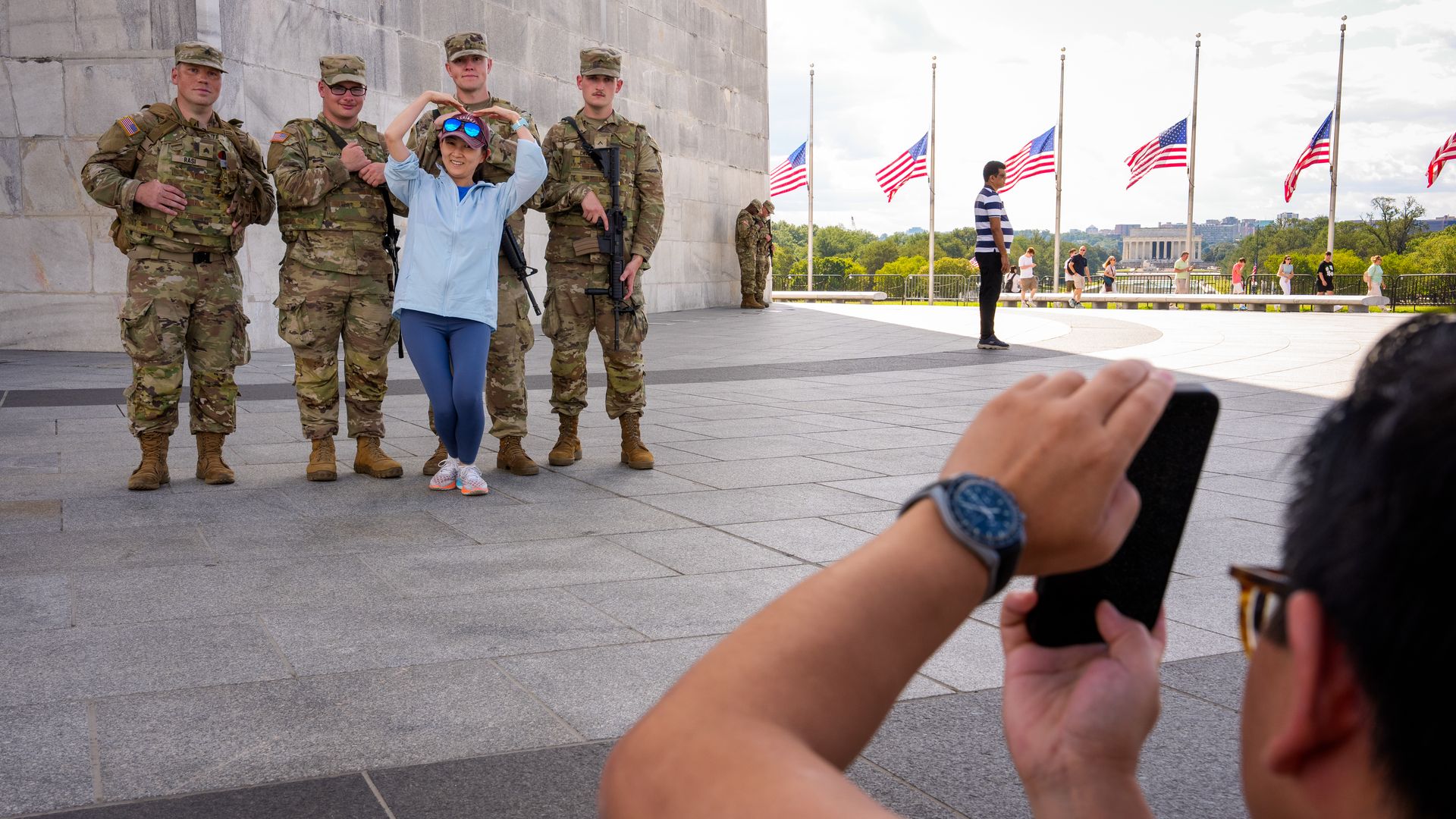 A woman in blue poses making a heart with her arms in front of four National Guard members in camouflage gear. A man in foreground takes their photo near American flags at a monument.