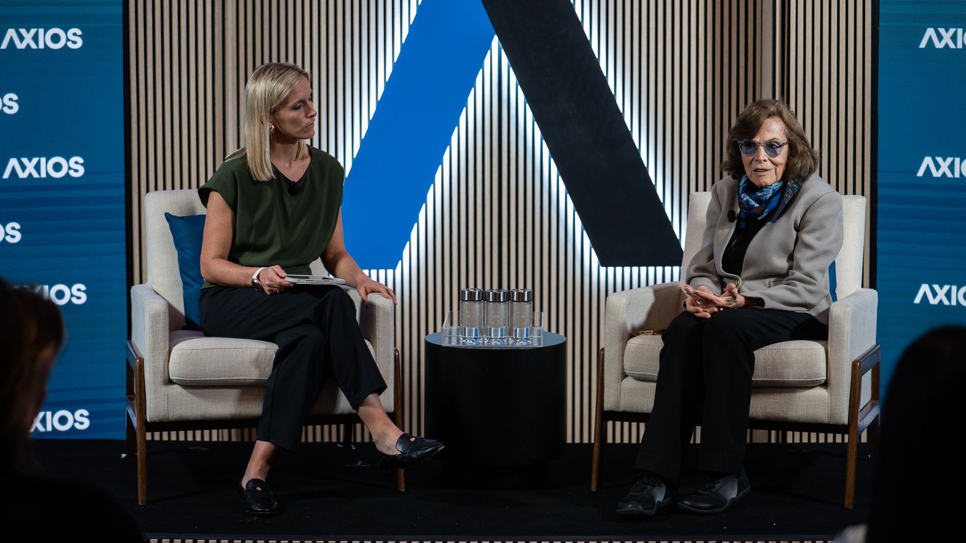 Two women sit on a stage in beige chairs, talking. Blue Axios banners flank them; a large blue-black angled backdrop and a glass table with water between them.