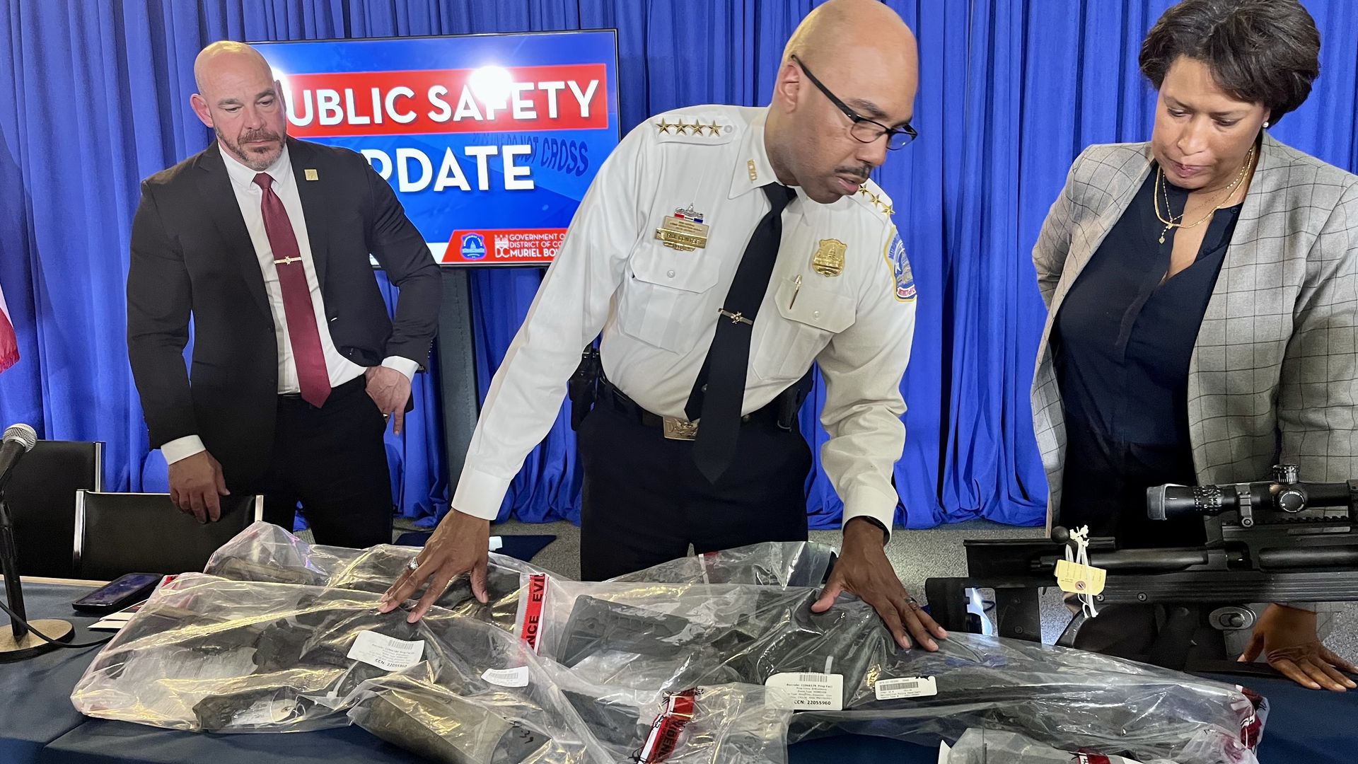 Police Chief Robert Contee stands before a table of six guns, in police bags, recovered from a suspected sniper