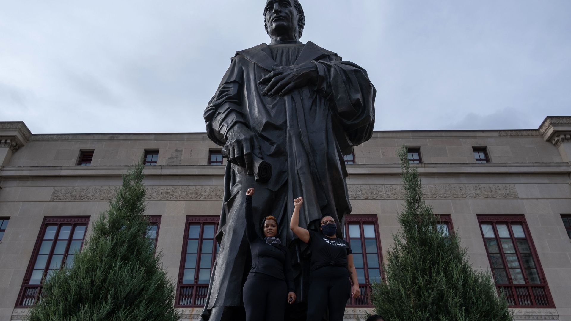 Protestors raise their fists in front of a Christopher Columbus statue on the City Hall courtyard. 
