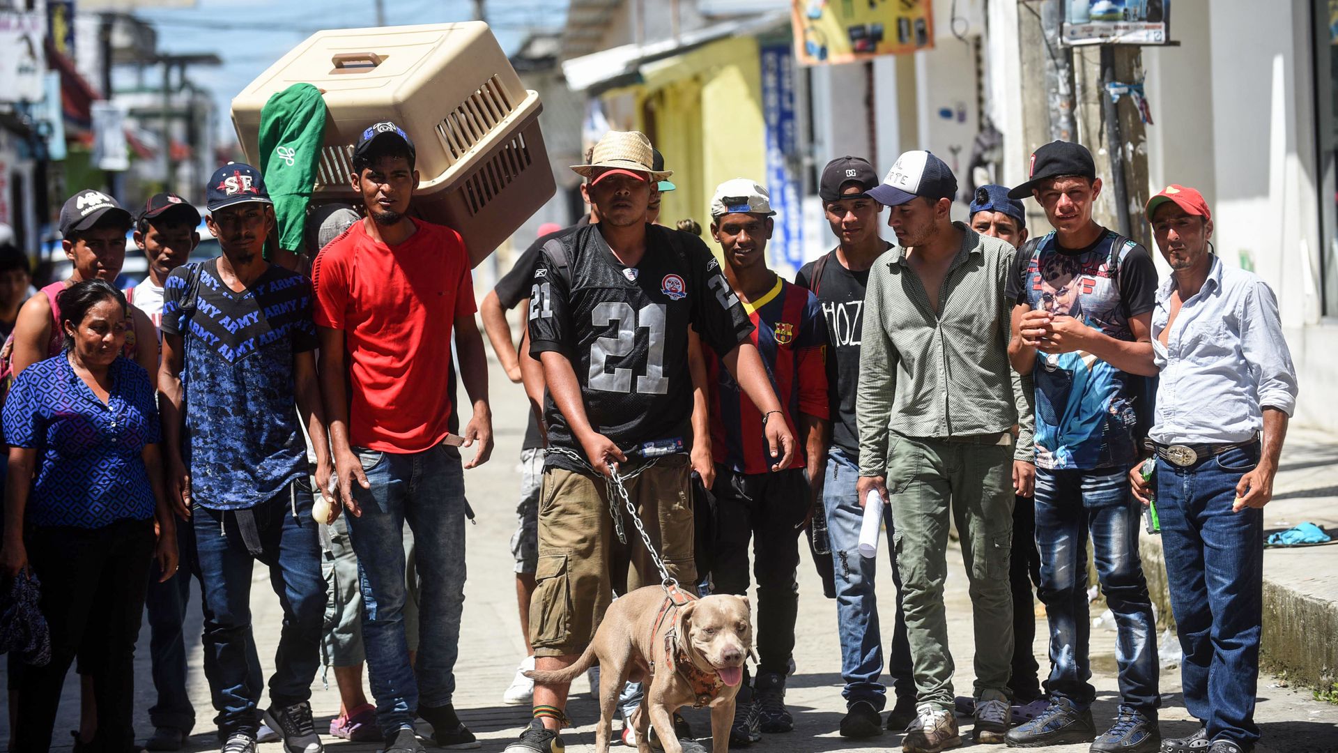 Honduran migrants stand in a row in a dusty road. One is holding the leash to a dog and another holds a dog cage over his shoulder. 