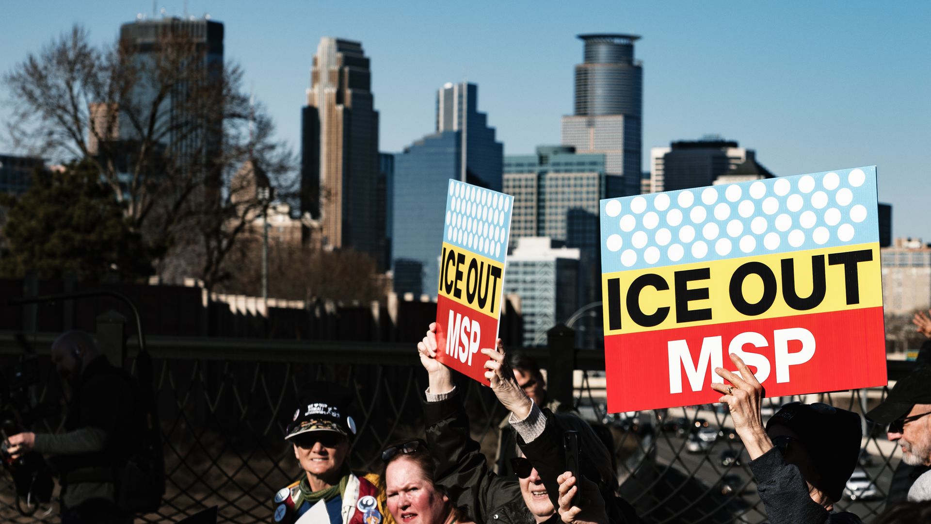 Group of people holding colorful signs reading "ICE OUT MSP" during a protest in front of a city skyline under a clear blue sky.