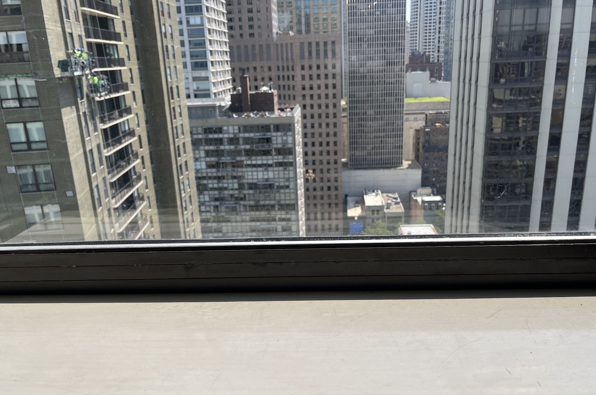Urban cityscape seen from a high window: tall beige and glass skyscrapers fill the frame, with a window sill at the bottom; clear daylight highlights the dense downtown skyline.