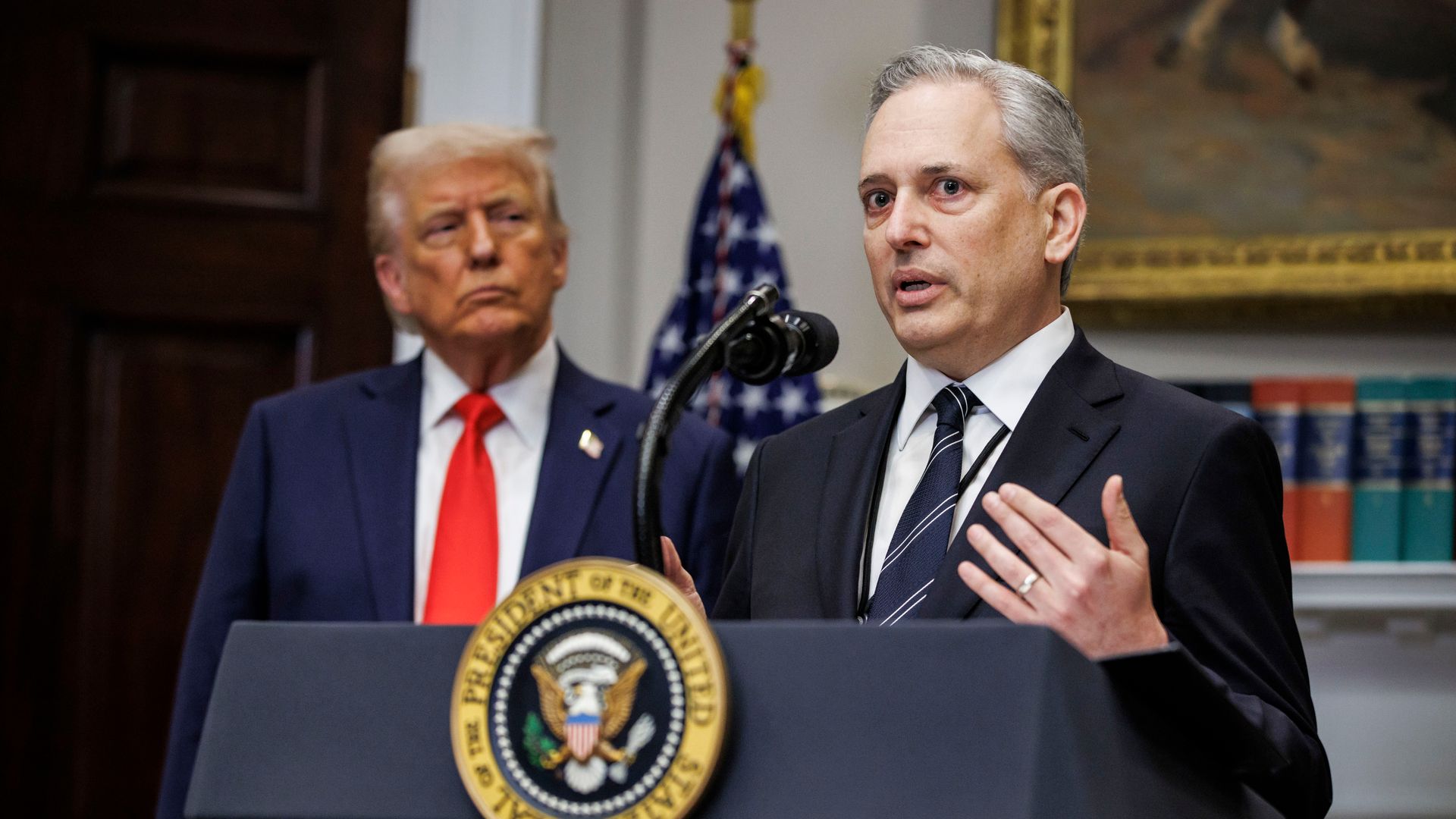 One man speaking at a podium while President Trump watches in the background, with a red tie on.