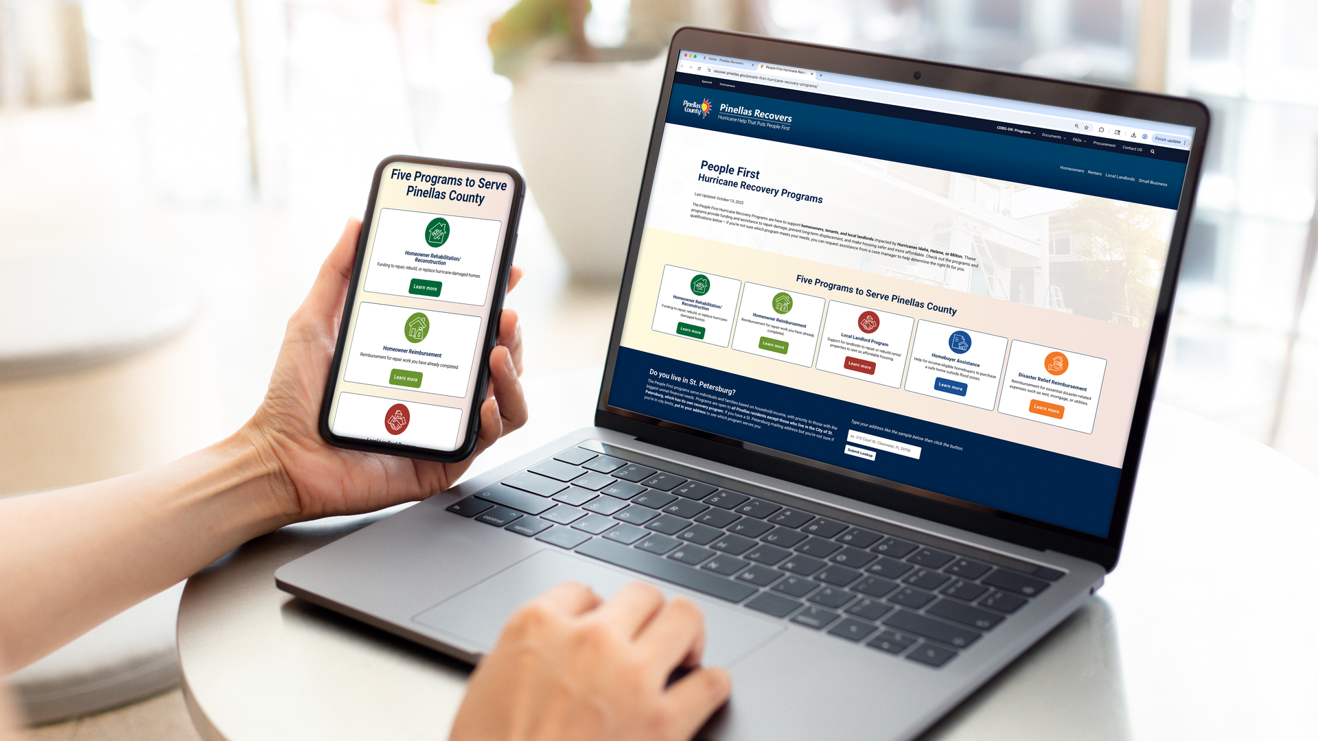 A hand holds a mobile phone while the other hand moves across a trackpad on a laptop. The mobile phone and laptop screens show websites that read "Pinellas Recovers, People First Hurricane Recovery Programs" and "Five programs to serve Pinellas County."