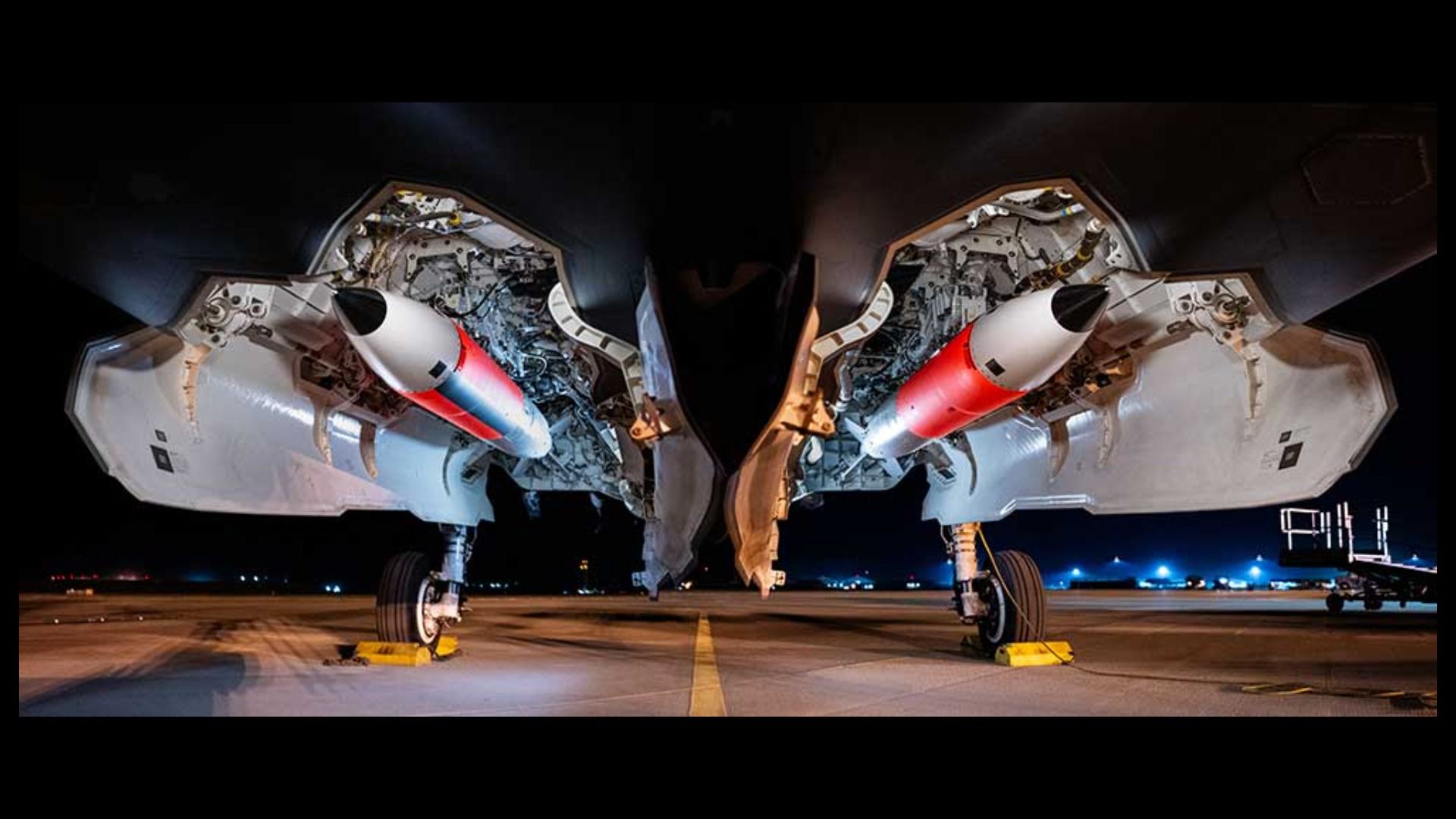 Underneath view of a fighter jet's open weapon bays showing two white and red weapons on a night runway with blue lights in the distance and yellow wheel chocks securing the aircraft.