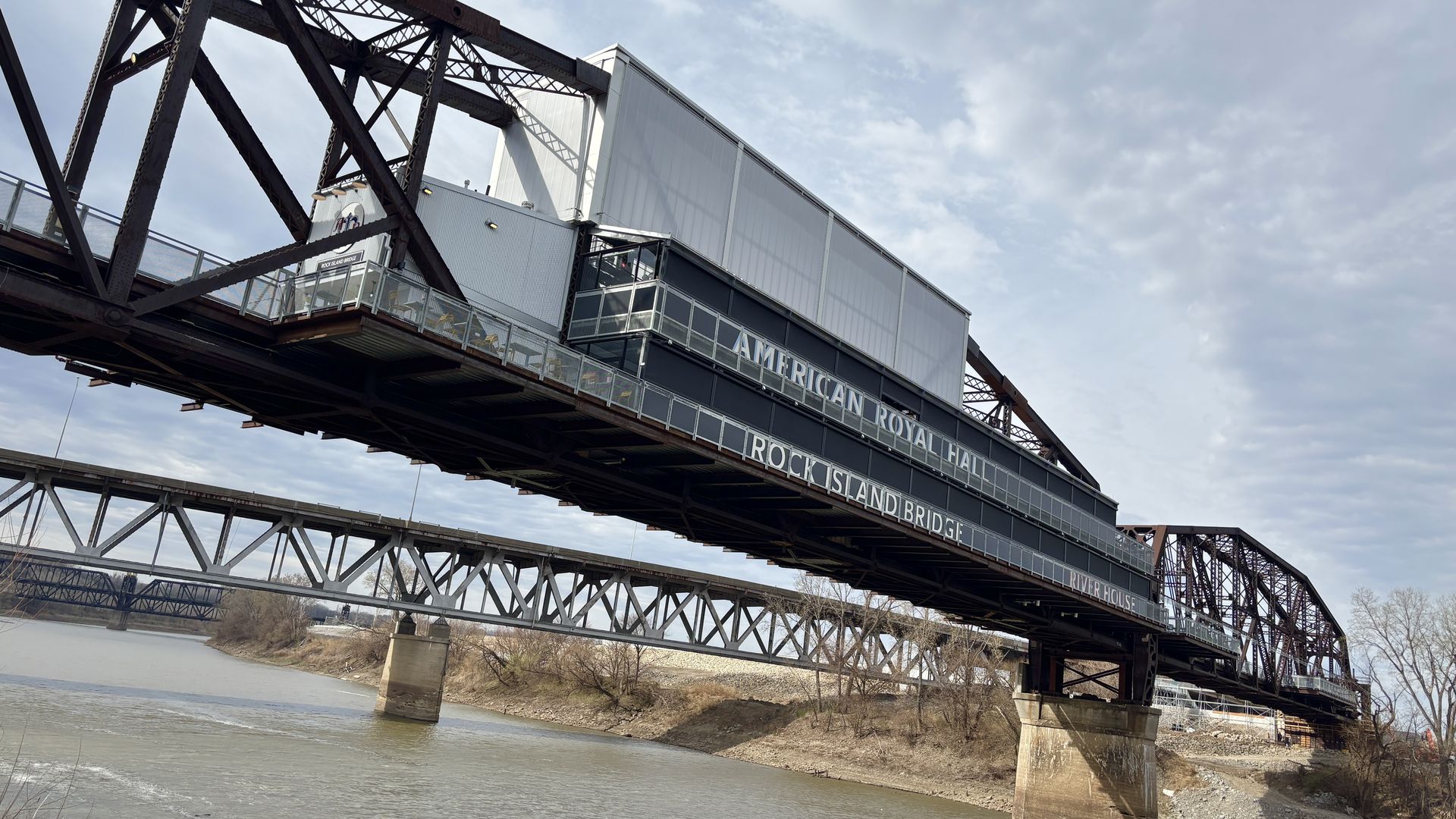 Dark steel swing bridge spans a calm river; a raised central section bears the words "AMERICAN ROYAL HALL ROCK ISLAND BRIDGE". In the background, a second truss bridge under a cloudy sky, with leafless trees along the shore.