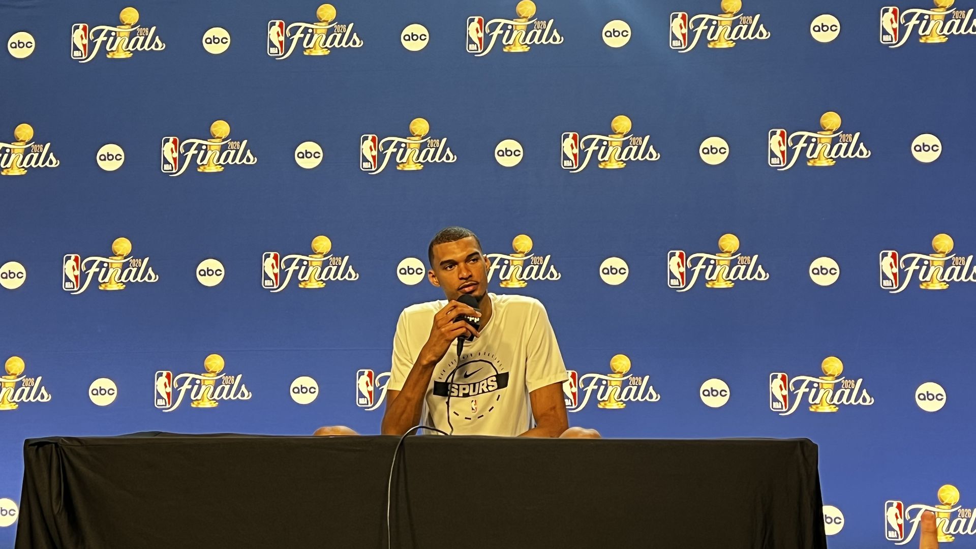 A man in a Spurs t-shirt sits at a press conference table, speaking into a microphone, in front of a blue backdrop covered with NBA Finals logos and ABC sponsor circles.