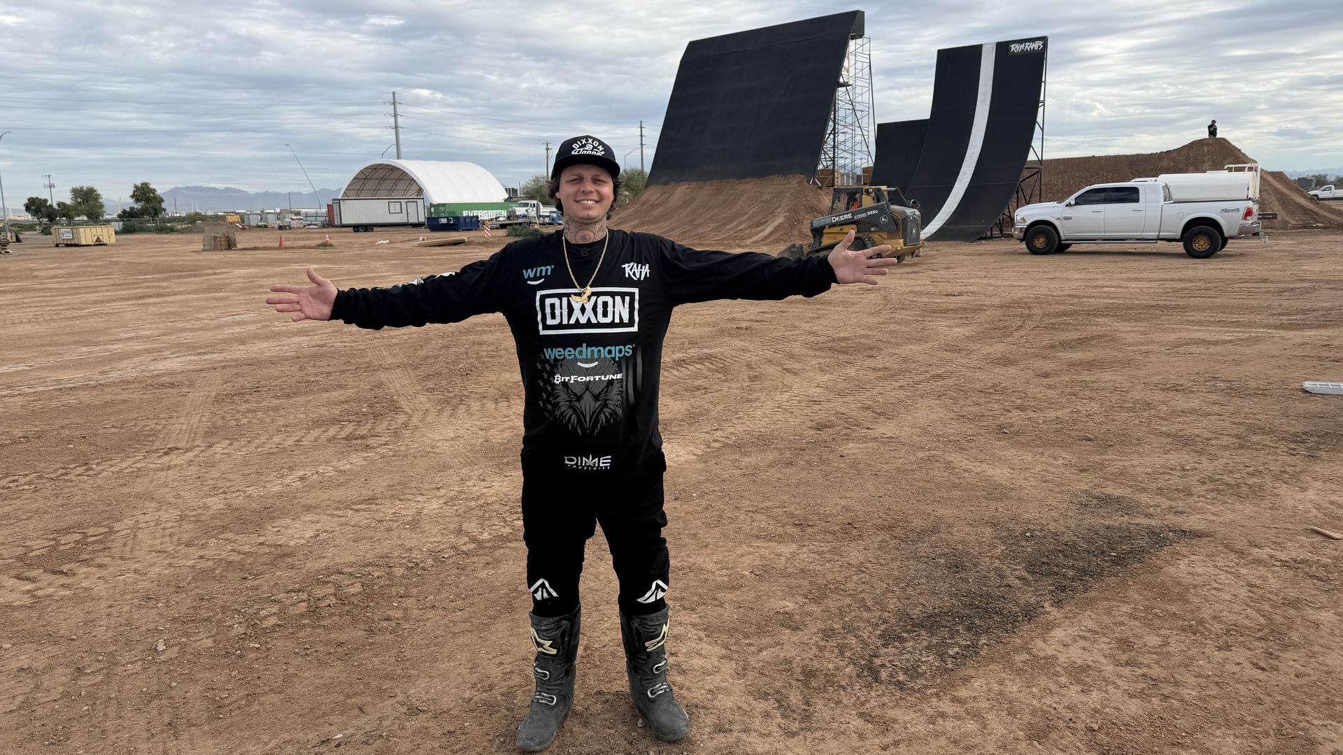 Person wearing black motocross gear and boots standing with arms wide open on a dirt field with large motocross ramp and white truck in the background under cloudy sky.