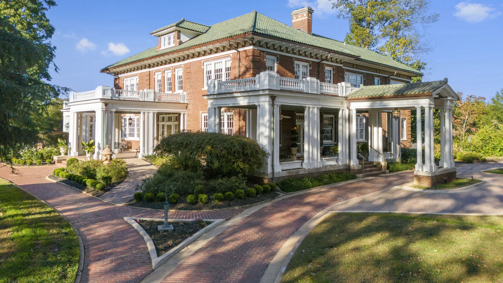 Brick mansion with a green tiled roof and white-columned veranda, surrounded by manicured shrubs. A curved brick driveway leads to the entrance on a sunny day.