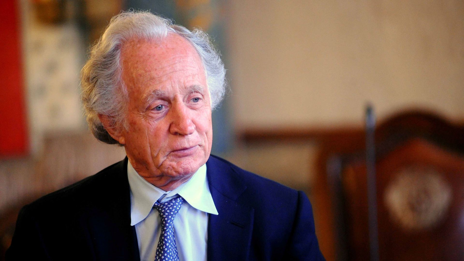 A man with white hair, a suit and a blue necktie looks to the side.