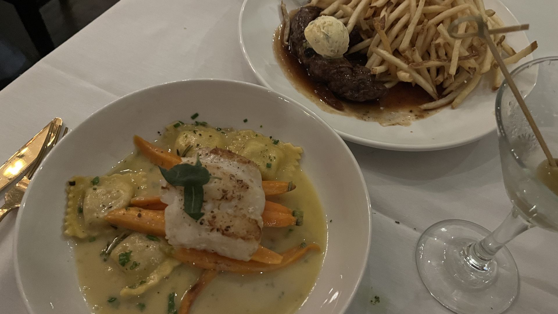Plate features a large mound of golden French fries atop a seared steak in brown sauce, topped with a scoop of mashed potato; a second plate with ravioli in yellow sauce sits nearby on a white table.