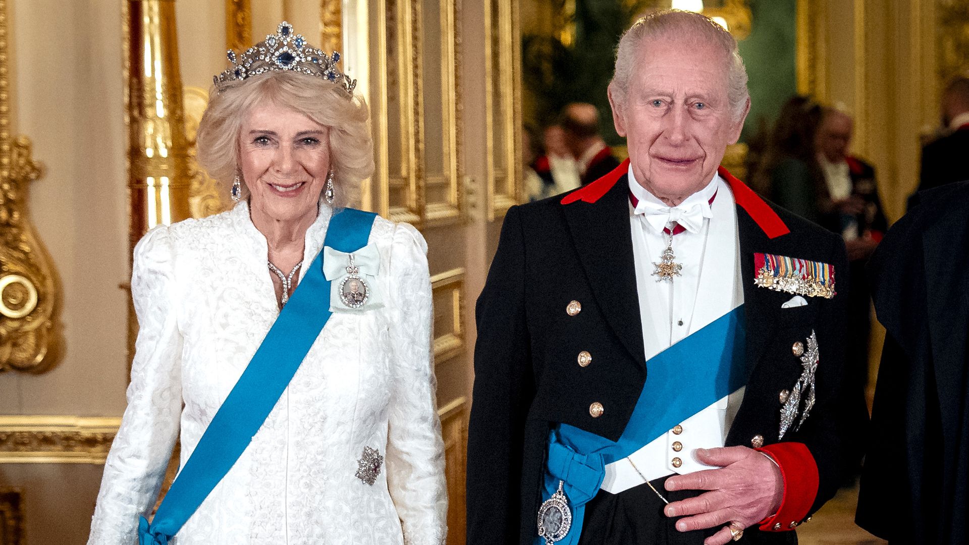 An elderly royal couple in a gilded hallway. The woman wears a white lace dress, a tiara, and a blue sash with a brooch; the man wears a formal black uniform with medals and a blue sash.